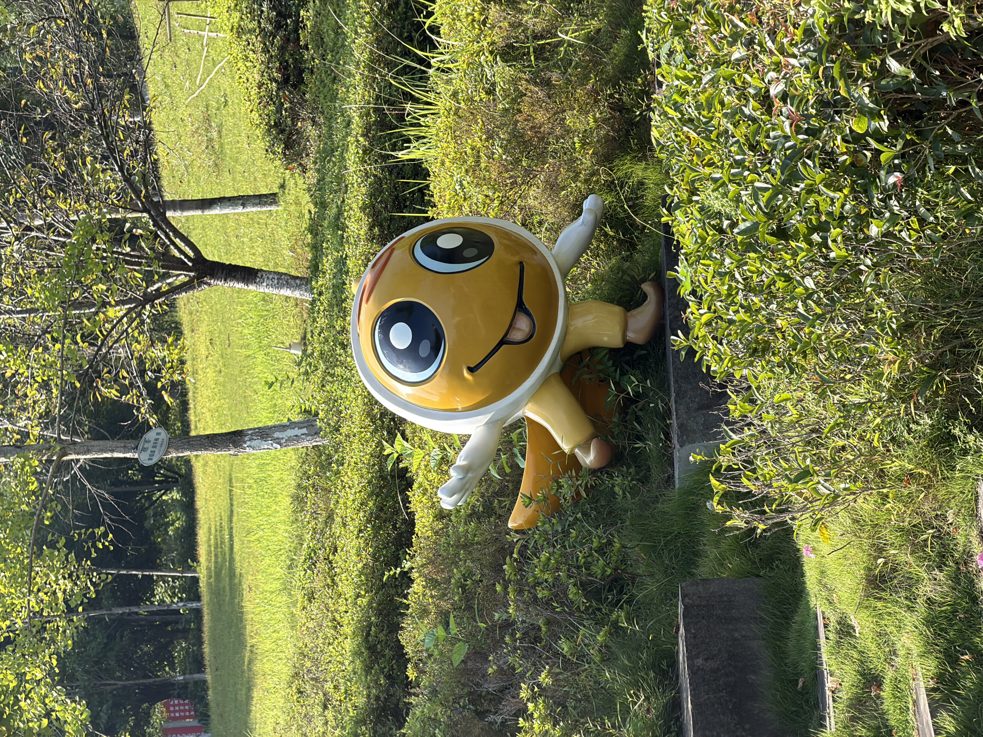 A yellow cartoon character statue with large blue eyes and an open mouth is situated on a grassy slope in a park. The statue is wearing a white helmet and has light brown legs and feet. It appears to be in motion, with one arm extended to the side. The background features several trees with green leaves and a lush green lawn. The scene is illuminated by sunlight, creating shadows and highlighting the vibrant colors of the vegetation and the statue. The location is identified as Jiangjin District, Chongqing, China, specifically within a park.