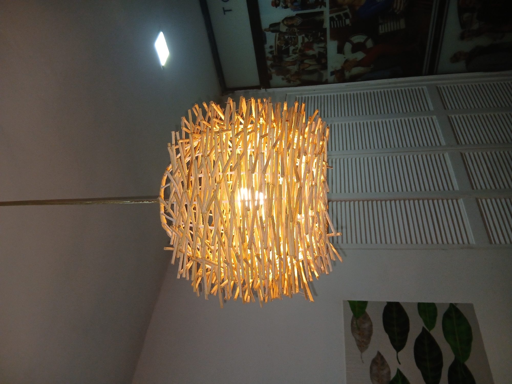 A low-angle shot of a decorative, cylindrical light fixture comprised of numerous small wooden sticks interwoven together. The light fixture hangs from the ceiling by a thin rod. The room has white walls, a white paneled ceiling, and a wall hanging depicting leaves. The location is in Playa El Agua, Venezuela.