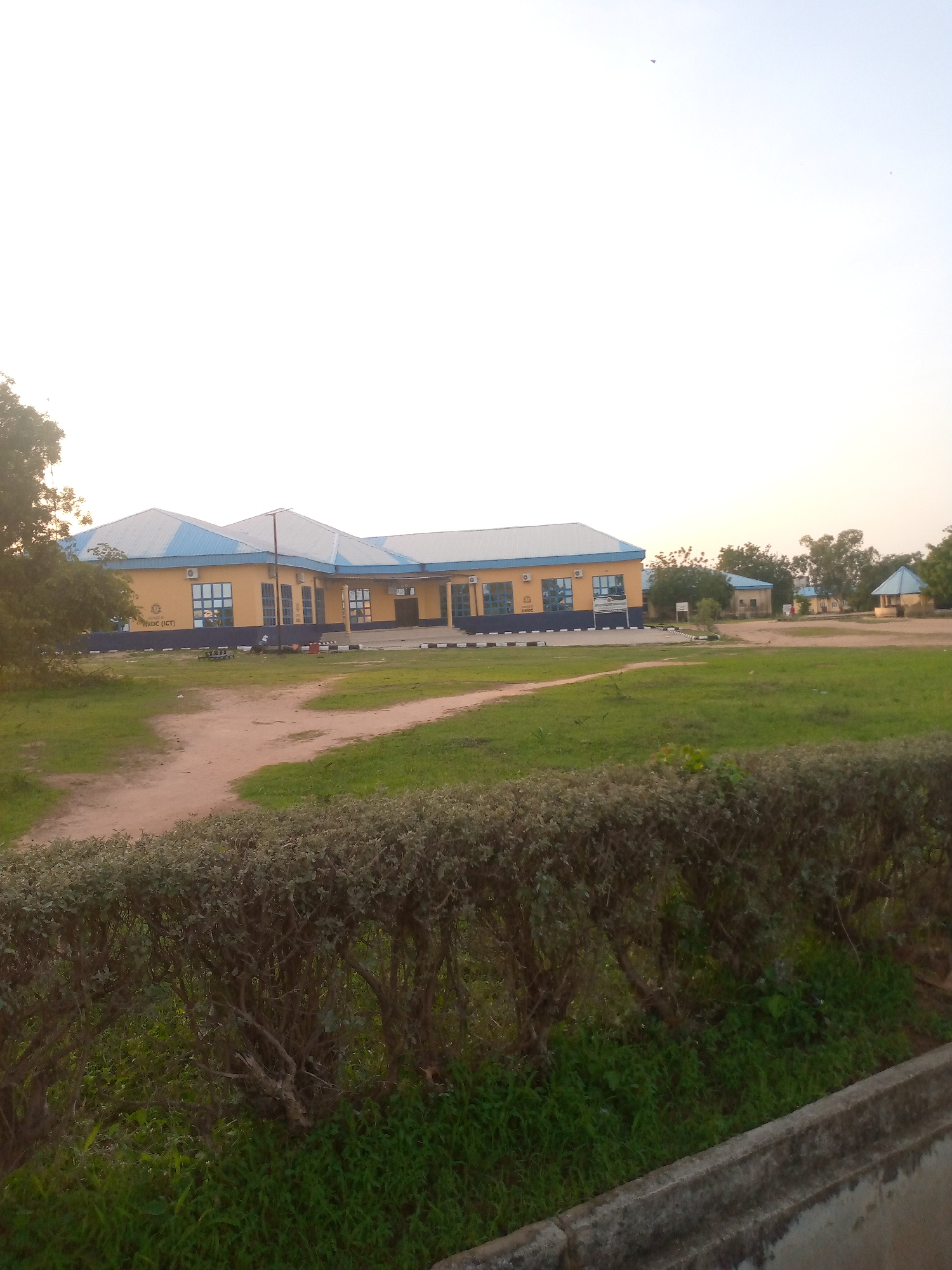 The image depicts a building with a yellow facade and blue roof, located in Jalingo, Nigeria. The building appears to be a public institution, possibly an educational or administrative center, with several windows and an entrance. The surrounding area is characterized by a grassy field, a dirt path, and some trees and bushes. The sky is clear and bright, suggesting daylight. There are no people visible in the image. The scene conveys a sense of openness and a rural or suburban setting within Jalingo.