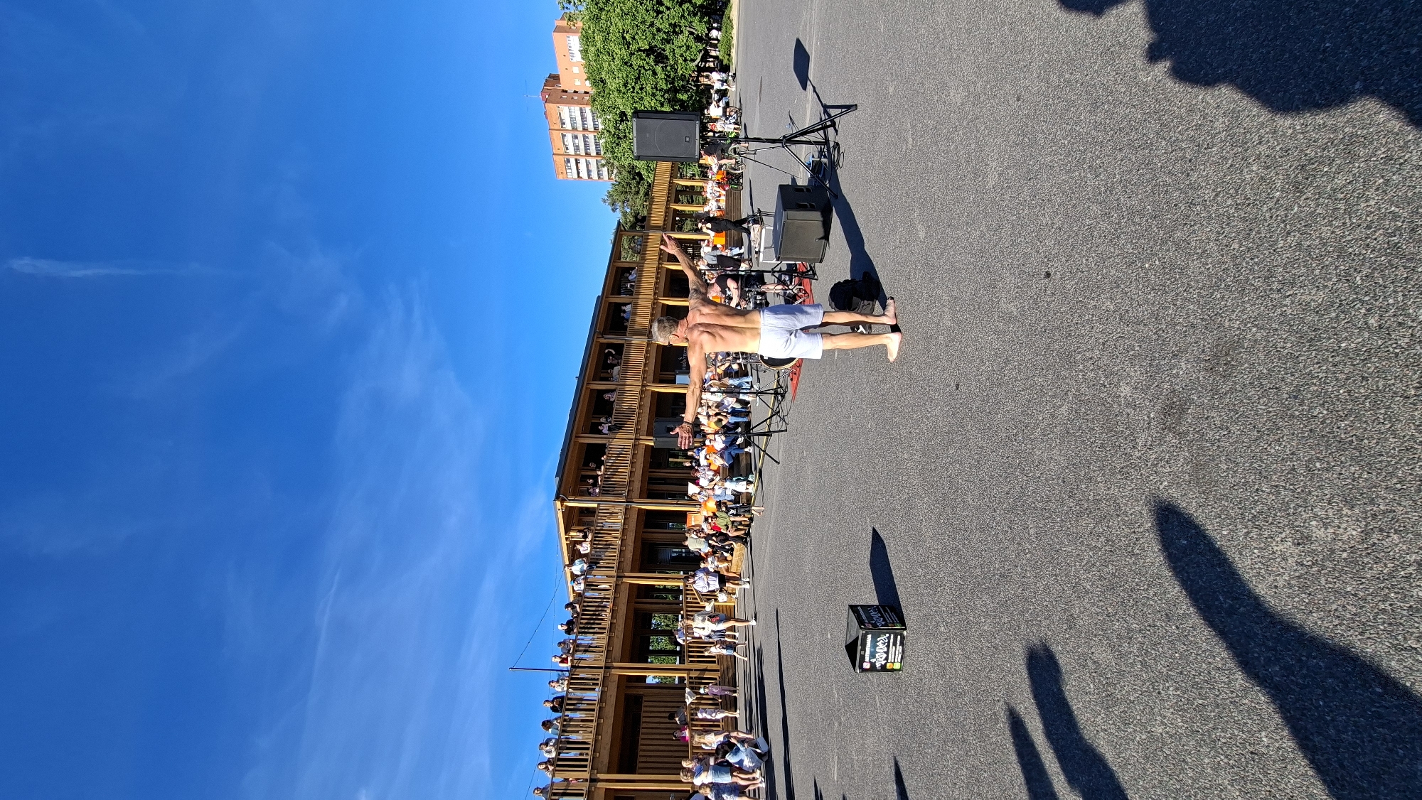 The image depicts an outdoor event on a sunny day, likely in Tallinn, Estonia. In the foreground, a man is standing shirtless with his arms outstretched, facing away from the camera. He is barefoot and wearing white shorts. Behind him, an audience is seated and standing, observing the event. A wooden building with multiple levels and balconies, filled with people, is visible in the background. There are also trees, two multi-story buildings, and a clear blue sky. Equipment such as speakers and stands are present, suggesting a performance or presentation is taking place. The ground is paved, and shadows of people are cast across it, indicating the direction of the sun. The overall scene suggests a public gathering in an urban or semi-urban setting.