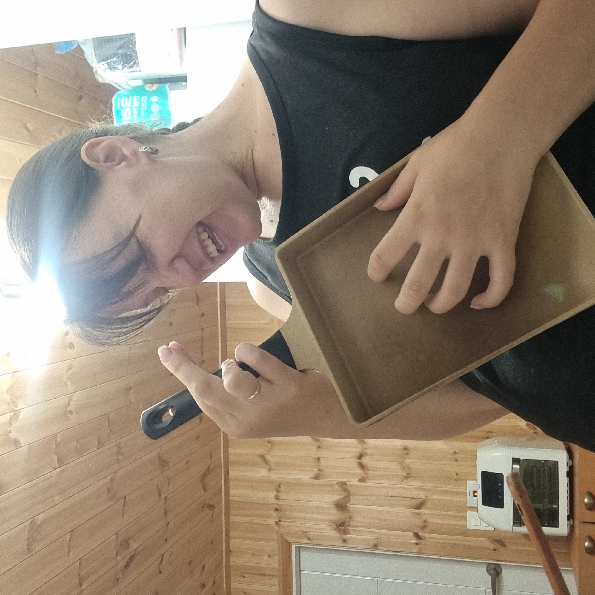 In an indoor setting with wood-paneled walls and ceiling, a person is holding a rectangular object resembling a baking pan, with their left hand positioned on it and their right hand holding the handle of a spatula-like utensil. The person's mouth is open in a laugh or shout, and their eyes are closed or squinting, illuminated by a bright light source reflecting off their forehead. They are wearing a black sleeveless shirt and a ring on their left ring finger. In the background, an air fryer and what appears to be a roll of toilet paper are visible. The scene suggests a playful or humorous moment.