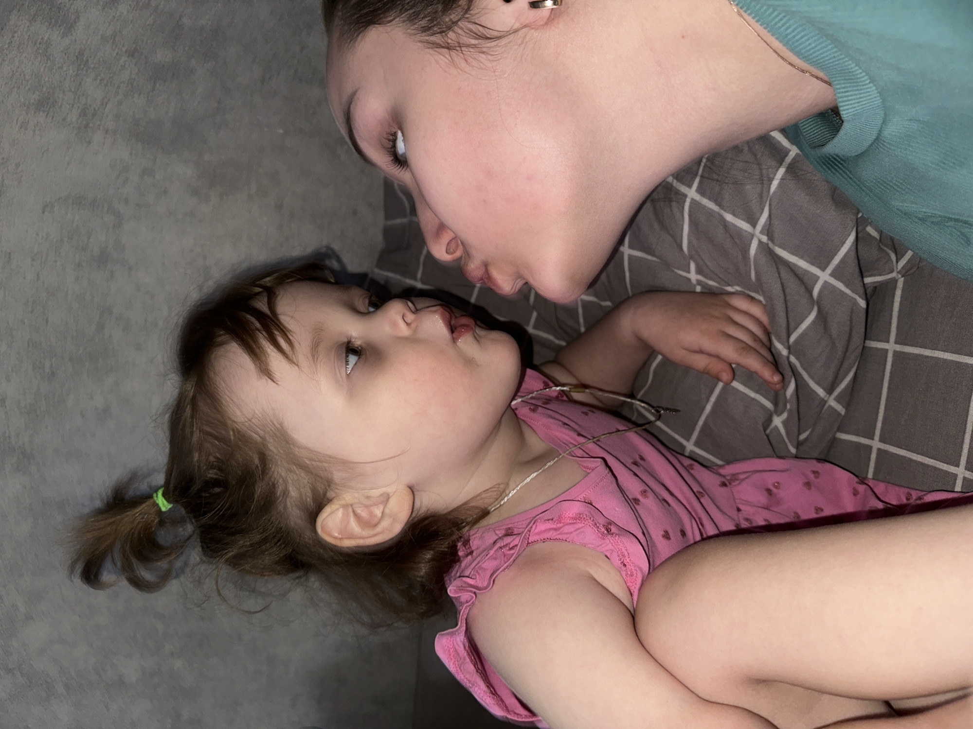 A person is lying on a gray, checkered pillow and attempting to kiss a child who is lying next to them. The child is wearing a pink shirt with small hearts on it and a necklace, and has their hair tied in a small ponytail with a green elastic band. The person attempting to kiss the child is wearing a teal shirt and an earring in their left ear. The background is a gray wall. The location is not discernible from the image.