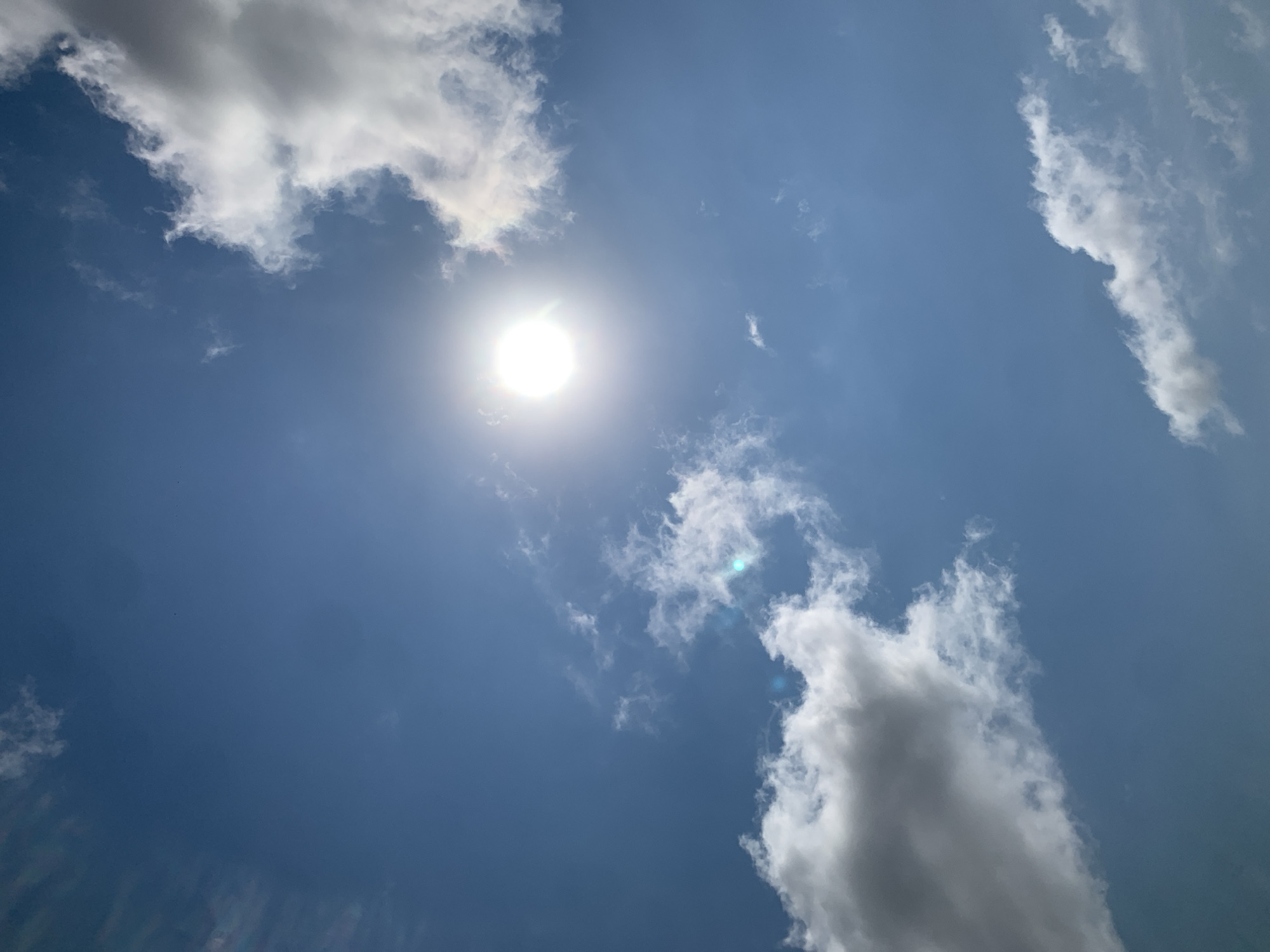 The image shows a bright sun high in a clear blue sky with scattered white clouds. The sun is slightly off-center to the left, with a bright glare. Several wispy clouds are visible, some more dense than others, in various parts of the sky. The lighting suggests it is daytime, likely mid-morning or afternoon. No specific landmarks or indications of Khazar Raion, Azerbaijan are visible in the image. The scene is purely natural, focusing on the sky and celestial bodies.