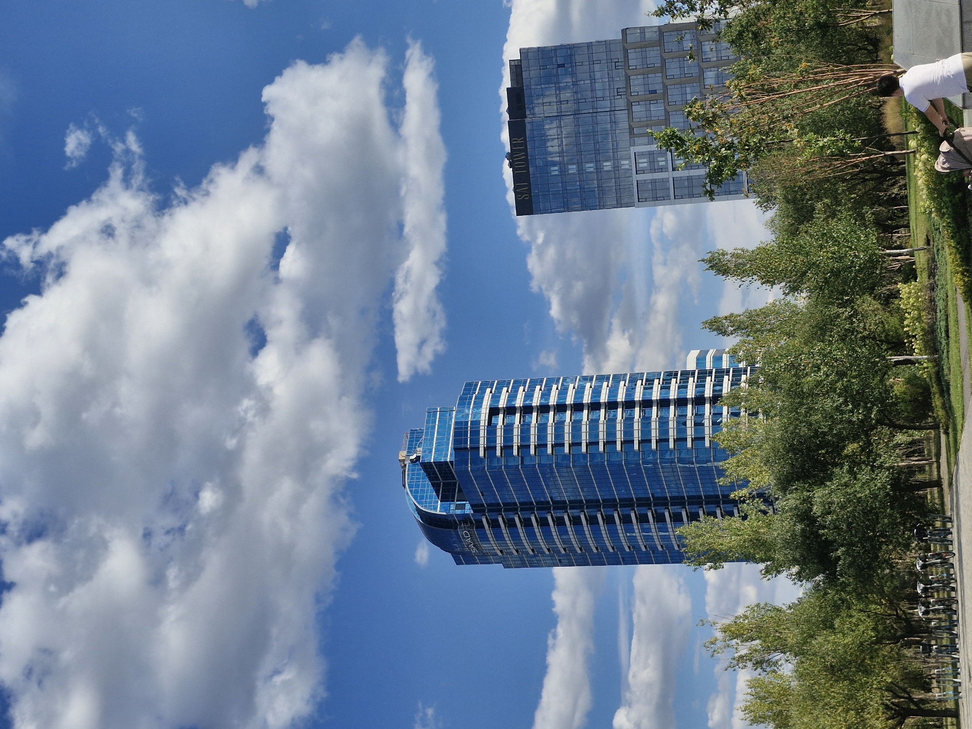 The image depicts a sunny day in Astana, Kazakhstan, with two modern skyscrapers dominating the background under a bright blue sky filled with fluffy white clouds. The skyscraper on the left is a tall, sleek building with a distinctive curved top, entirely clad in blue glass. The word "SAADE" is visible on its facade. To its right stands another skyscraper, also featuring a glass exterior, with the words "SAT TOWER" prominently displayed at the top.
In the foreground, a lush green park area is visible, with numerous trees of varying heights and foliage. A row of bicycles is parked in the middle ground, suggesting a public space or rental station. A person, dressed in a white t-shirt and light-colored shorts, is pushing a stroller along a path in the park, facing away from the camera. The overall scene conveys a sense of urban modernity blended with natural parkland.
