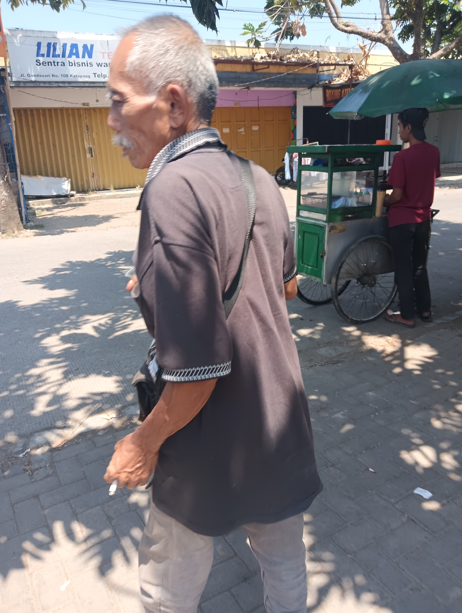 An elderly man with graying hair and a mustache, wearing a dark brown short-sleeved shirt and light brown pants, walks along a sidewalk in Katapang, Indonesia. He holds a cigarette in his right hand. In the background, a young man in a maroon t-shirt and black pants stands behind a green mobile food cart. The food cart has wheels and a green canopy. Behind the food cart are buildings with yellow doors and a sign that reads "LILIAN Sentra bisnis war". The ground is paved with interlocking concrete blocks, and shadows from trees are cast on the ground. The sky is visible and appears clear.
