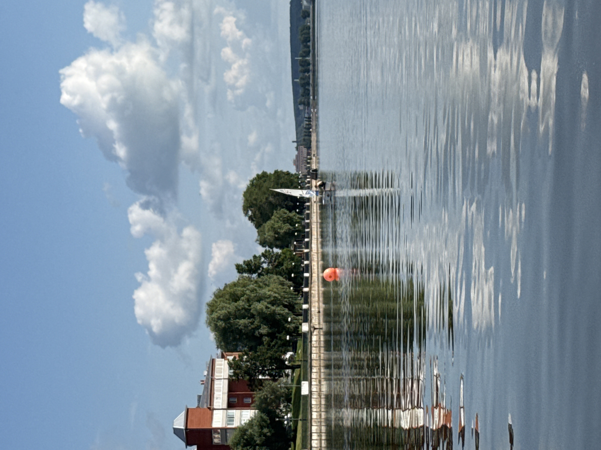 This image depicts a serene waterfront scene in Votkinsk, Russia. The foreground is dominated by the calm, rippling water of a lake or river, reflecting the sky and surrounding structures. The reflection shows textured patterns created by the movement of the water.
In the middle ground, a red brick building with white trim and a distinctive grey-capped tower stands prominently on the left bank. This building appears to be an institutional or residential complex. Adjacent to it are lush green trees lining the waterfront promenade. Several lampposts are visible along the embankment, suggesting a well-maintained public space.
Further out on the water, a small sailboat with a white sail is visible. A single person is steering the boat, indicating recreational activity. Off to the right of the sailboat, an orange spherical buoy floats on the water's surface.
The background shows a distant treeline and some industrial structures or city buildings along a gentle incline under a bright blue sky with scattered white clouds. The overall impression is one of tranquility and a pleasant day by the water. The location is identifiable as Votkinsk, Russia, likely along the Kama River or one of its tributaries, given the characteristic landscape and architectural style.