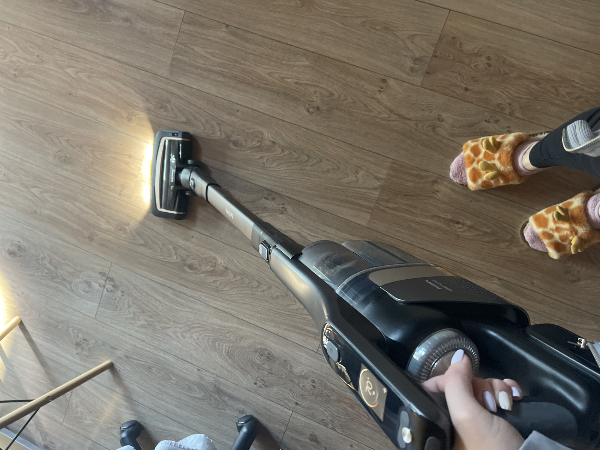 An indoor, eye-level shot depicts a woman vacuuming a wood floor with a black Philips cordless vacuum cleaner. The vacuum's head is emitting a bright light. The woman's hand, with light-colored nail polish, is visible on the vacuum's handle. She is wearing giraffe-patterned slippers. A partially visible chair leg is in the lower-left corner. The location is likely a private residence in an urban area; however, the specific location in Sofia, Bulgaria, cannot be determined from the image alone.