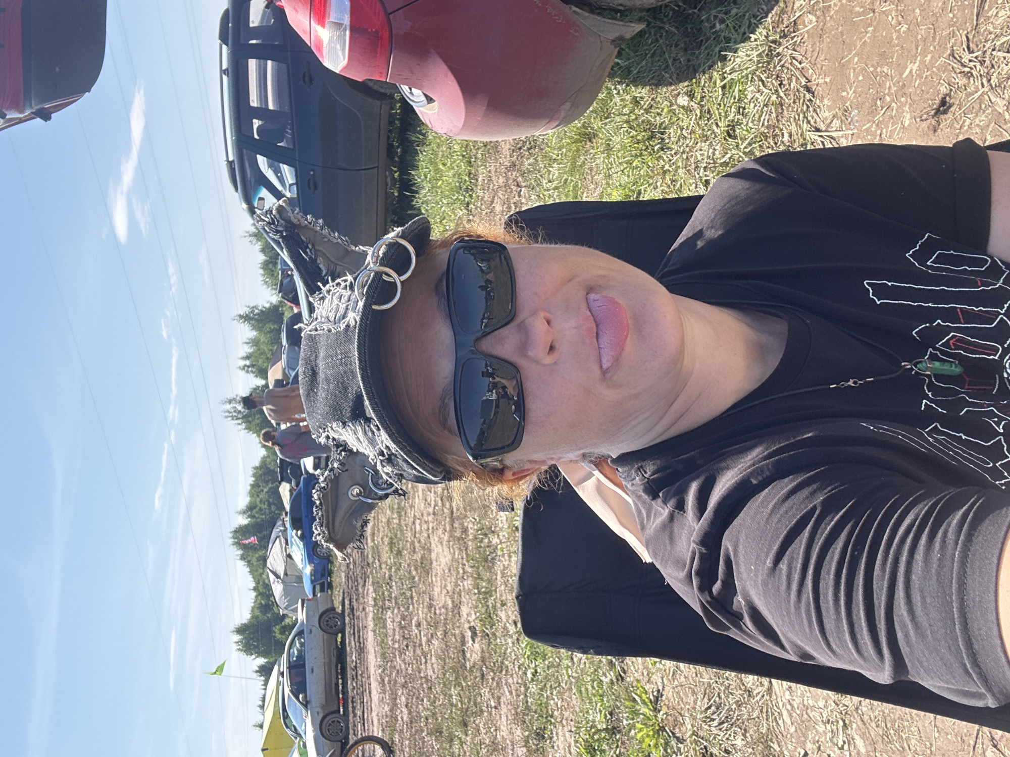 In the city of Chormoz, Russia, a person with reddish hair and wearing sunglasses and a black cap with distressed fabric and rings is sitting in a black chair. They are sticking their tongue out at the camera. The person is wearing a black t-shirt with a pendant necklace. In the background, there are several cars parked on a dirt and grass field, with trees and tents visible in the distance under a blue sky with some clouds. The scene appears to be a festival or outdoor gathering.