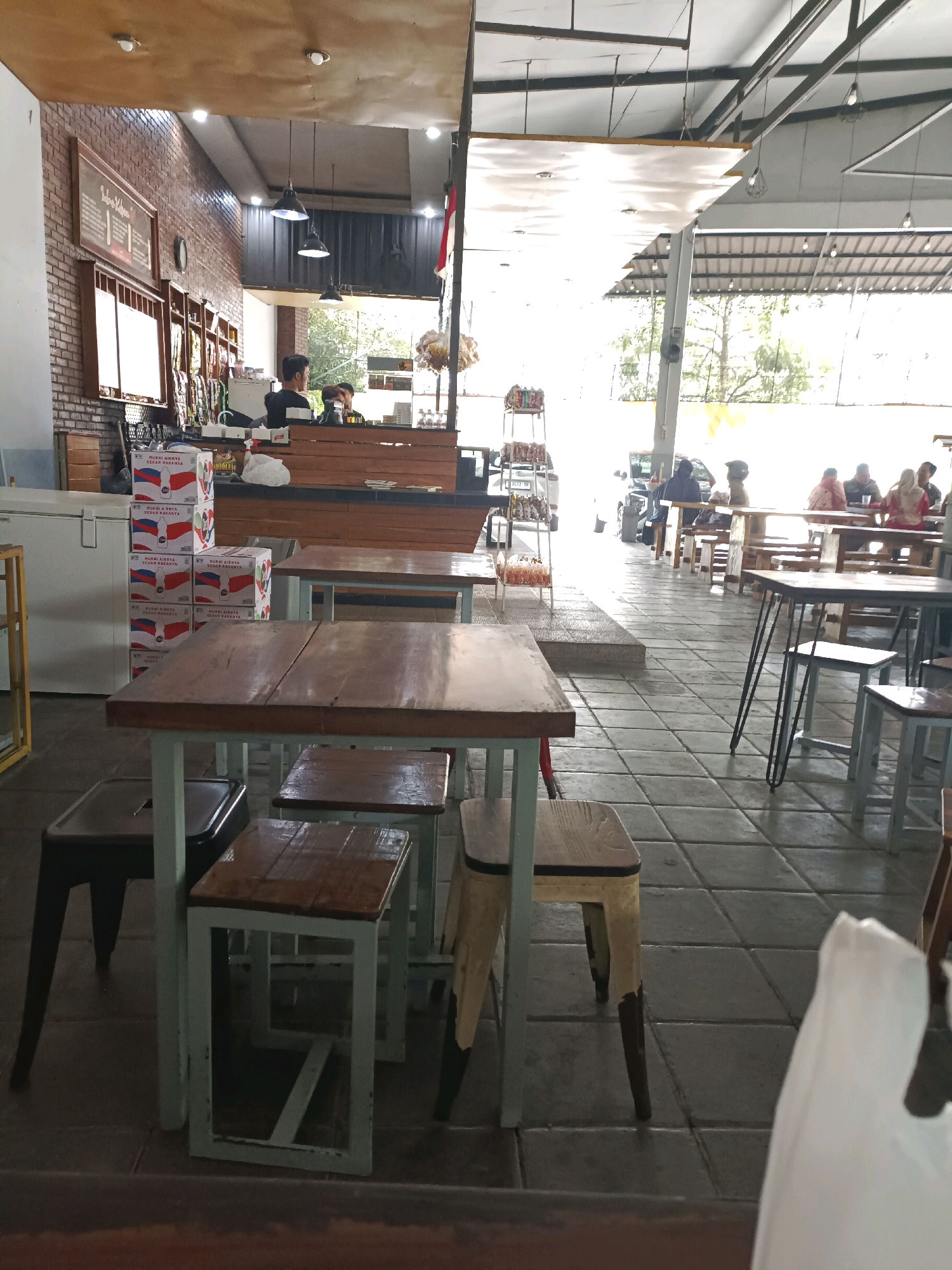 The image depicts the interior of a cafe in Made, Indonesia. In the foreground, several wooden tables with metal legs are arranged, some with attached benches or stools. Stacks of boxes, possibly containing beverages, are visible to the left of the tables. A counter area with a wooden facade is situated in the midground, where two individuals, presumably staff members, are interacting. Behind the counter, shelving displays various items. The cafe's seating area extends into the background, with multiple tables and patrons seated. The overall ambiance suggests a casual dining or gathering space. The environment appears to be an open-air or semi-open-air setup, with natural light filtering in from the sides and overhead.