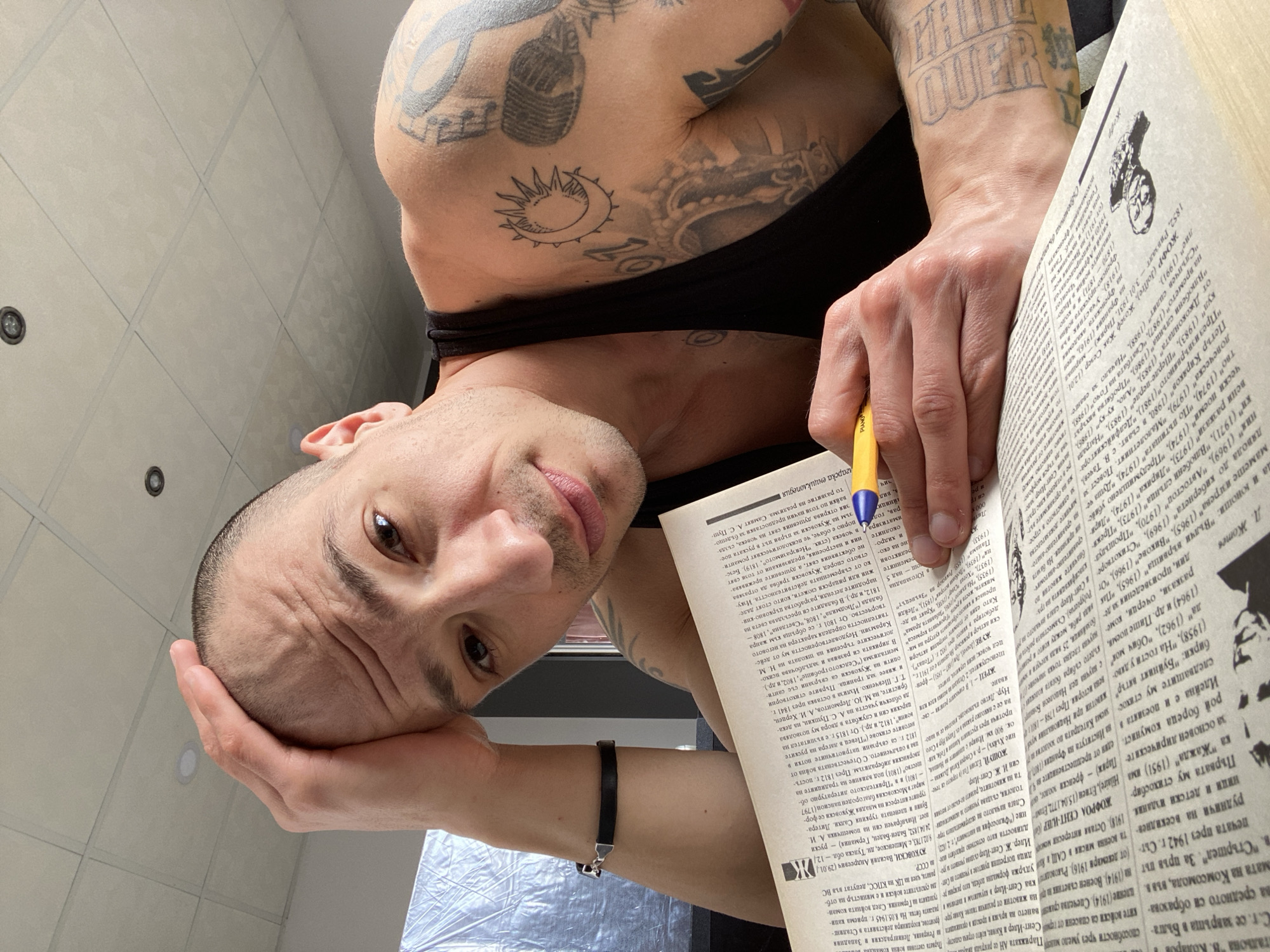 A man with tattoos on his arms is indoors, holding a book open with one hand and a pen in the other. His head is resting on his other hand. The book contains text in Cyrillic script, possibly relating to Bulgarian history or literature. The environment suggests an indoor setting with white tiled ceilings. No specific landmarks indicate the location is Pleven, Bulgaria.