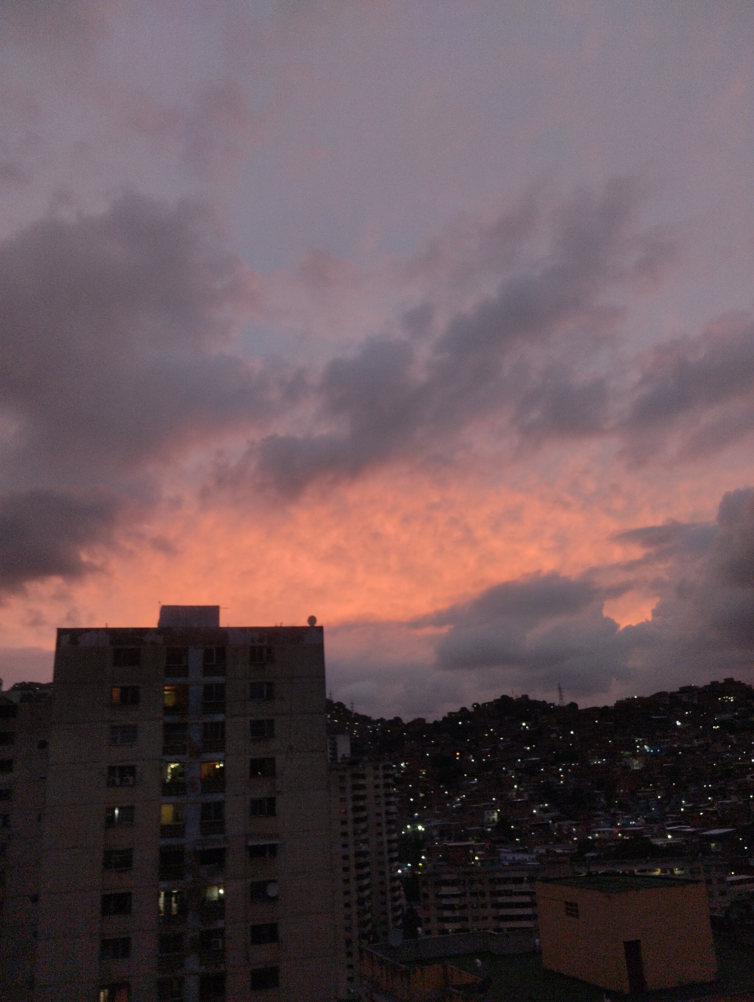 The image shows a cityscape in Caracas, Venezuela, at twilight. A tall, rectangular apartment building dominates the left side of the frame. The building is a drab gray color and features numerous windows, some of which are illuminated. In the background, a hillside is densely populated with houses and structures, their lights twinkling in the dusk. Above, a dramatic sky displays a gradient of color, transitioning from a fiery orange near the horizon to darker shades of gray and purple at the top. The clouds are scattered and textured, adding depth to the scene.