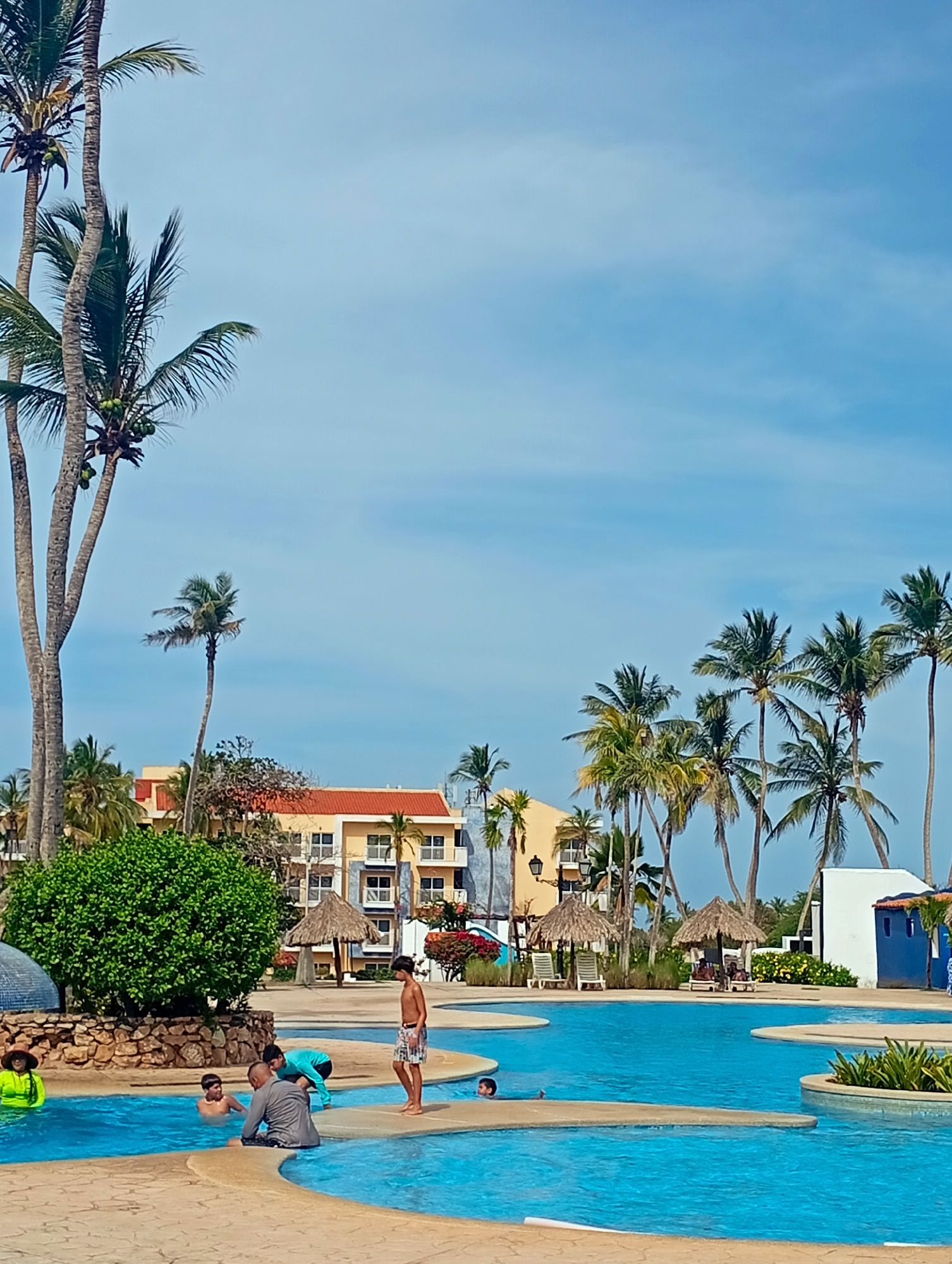 The image depicts a pool scene at a resort in Playa El Agua, Venezuela. Several individuals are in and around a multi-level swimming pool. Some are wading, some are swimming, and others are standing on the pool's perimeter. The pool has several small, shallow sections connected by narrow walkways.
Behind the pool are several palm trees, thatched-roof umbrellas, and lounge chairs. Further in the background, there's a multi-story building with an orange roof, indicative of a resort or hotel. The sky is light blue with scattered clouds. Vegetation is also visible throughout the scene.