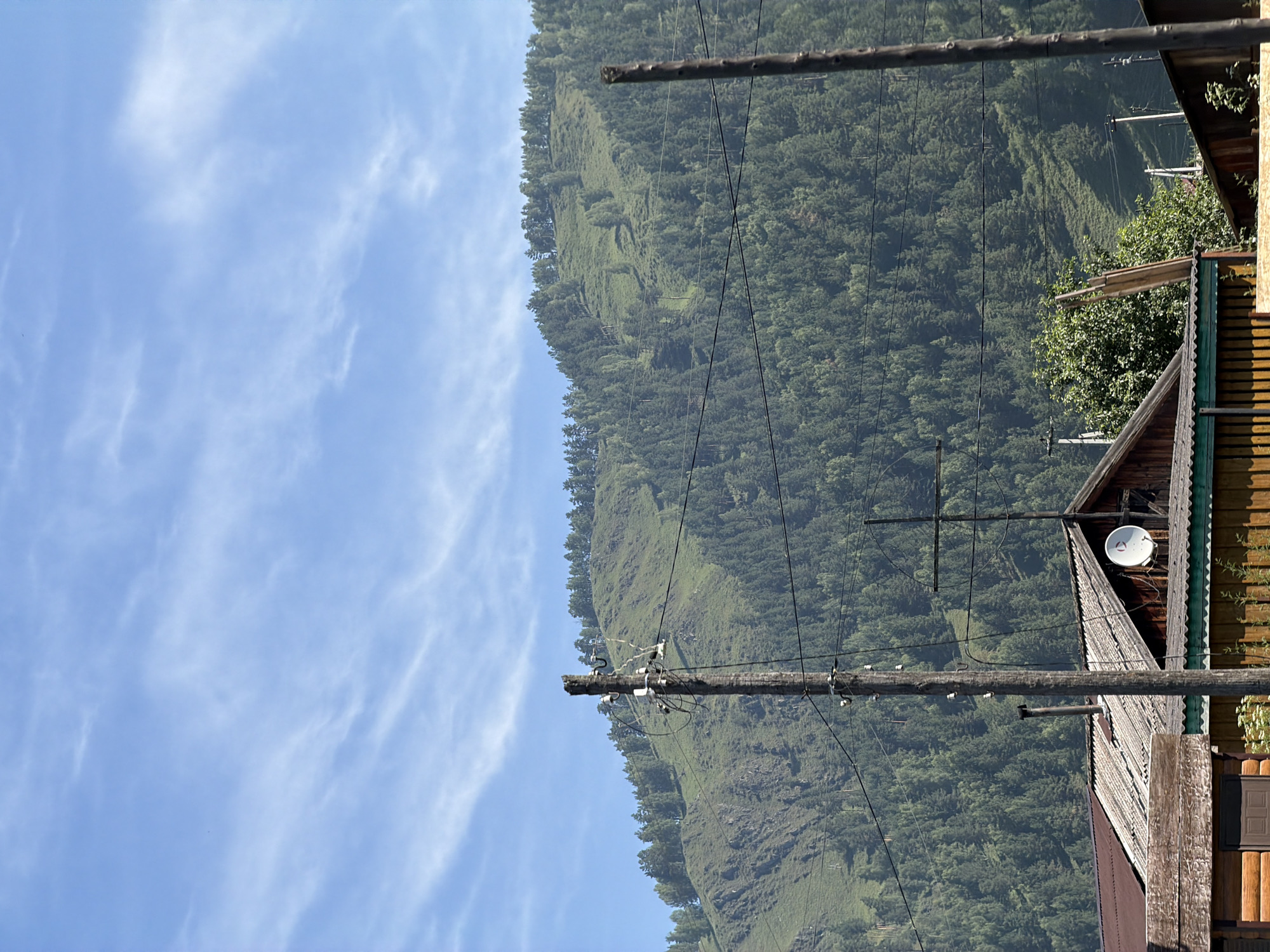 The image depicts a view of a rural settlement nestled at the base of a forested mountain under a partly cloudy blue sky. The foreground features several wooden structures, including houses with corrugated roofing and what appears to be a log cabin. Power lines are strung across the scene, supported by wooden utility poles. A satellite dish is visible on the roof of one of the buildings. The midground is dominated by a steep, verdant mountain slope covered in dense coniferous forest. The upper portion of the mountain displays sparser vegetation and rocky outcrops, with the sky above featuring wispy cirrus clouds. The location is identified as Elekmonar, Russia.