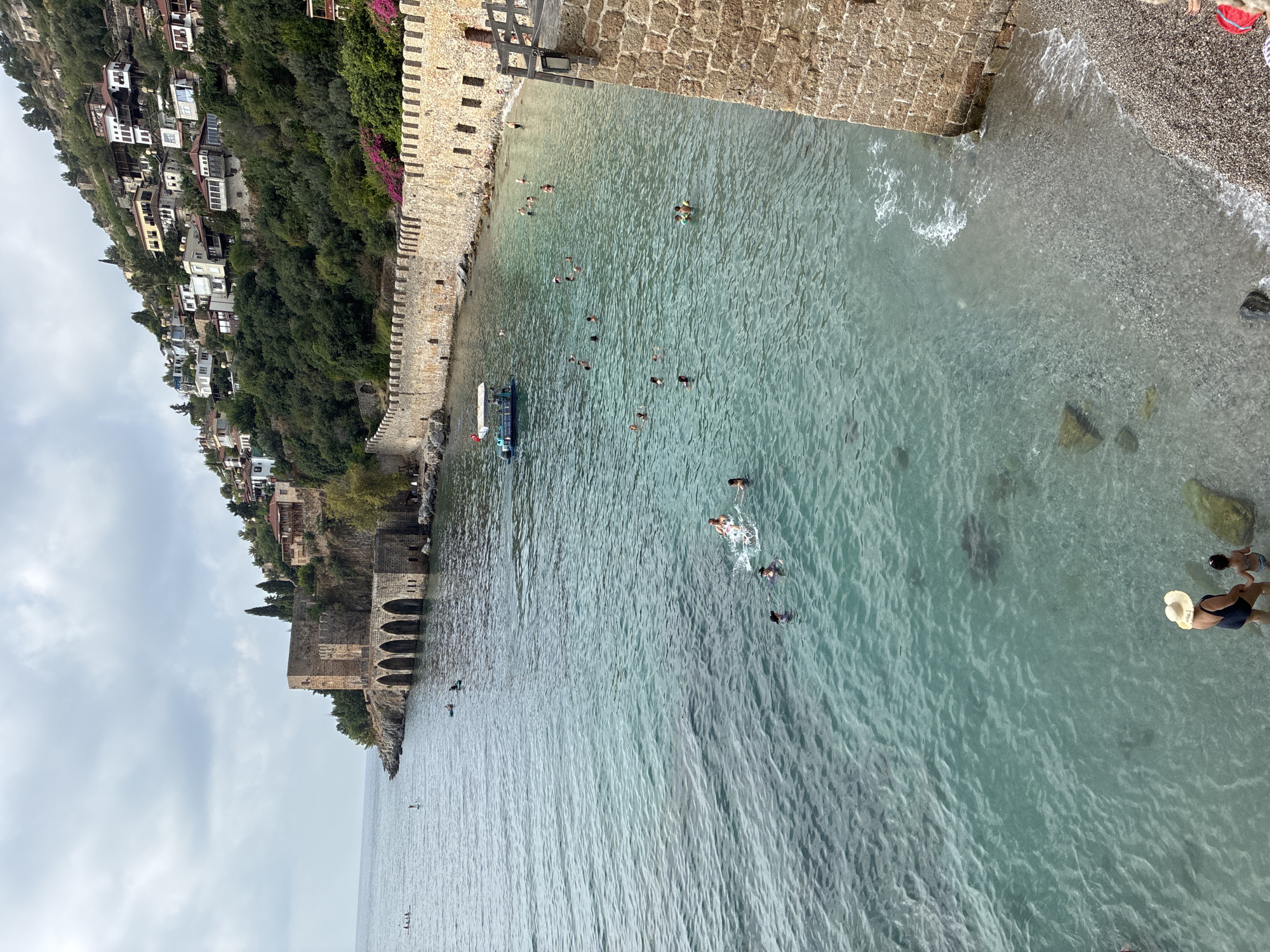 This image captures a picturesque view of the Alanya Castle walls and the Red Tower in Alanya, Turkey, overlooking a clear, turquoise sea filled with swimmers. The ancient stone fortifications, characterized by their arched openings and weathered texture, form a prominent barrier along the coastline. The Red Tower, a historical landmark, stands on a rocky outcrop to the left, its solid structure contrasting with the clustered white and red-roofed houses nestled on the steep hillside above.
The sea is populated by numerous individuals engaged in various water activities. Several people are swimming and playing in the water, while others are seen on paddleboards, enjoying the calm conditions. A small tour boat with an awning is navigating near the castle walls, adding a touch of modern tourism to the historical setting. The beach area, visible to the right and at the bottom of the frame, is covered in pebbles and small stones. Two people, a woman wearing a wide-brimmed hat and a child, stand at the edge of the water. The sky is overcast with scattered clouds, suggesting a temperate day. The overall scene conveys a sense of leisure and historical immersion in the vibrant coastal city of Alanya.
