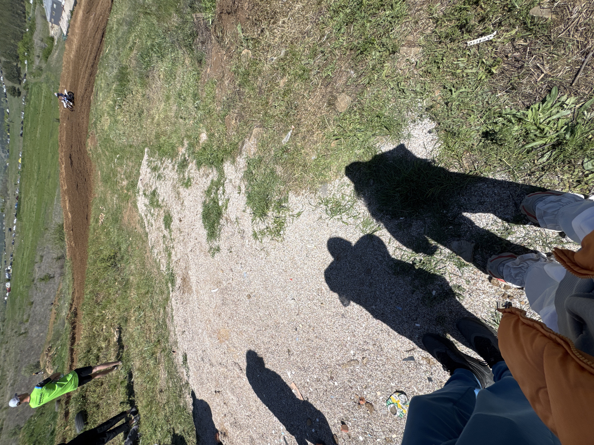 The image shows a dirt track with a motorcycle racer on the top left, a grassy area, and a gravel path with the shadows of people standing on it. The view is from above, looking down at the path. The shadow of a person wearing light-colored pants and shoes with an orange jacket is visible on the right. A person wearing a green shirt and a white hat stands on the grass to the left. The image appears to be taken in Belo Pole, Bulgaria, as indicated by the context.