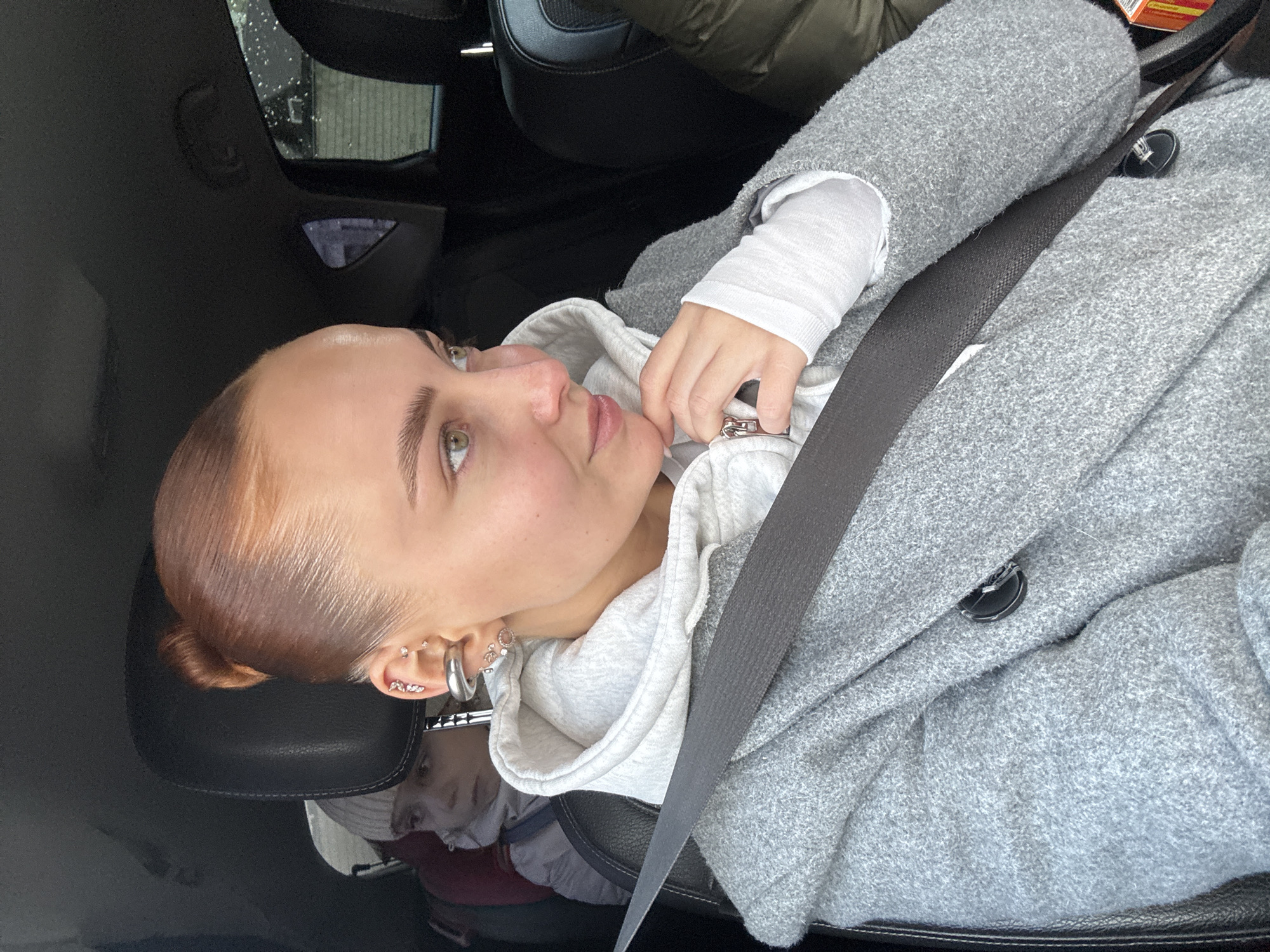 A young woman is depicted in the passenger seat of a car during the daytime in Chaikovsky, Russia. She is buckled in with a dark seatbelt that crosses her chest and lap. Her reddish-brown hair is neatly slicked back into a bun. She is dressed in a light grey hooded sweatshirt, layered under a textured grey coat or jacket, with a white long-sleeved top visible at her wrists. Her left ear showcases multiple piercings, including a prominent silver hoop, several small studs, and a curved bar adorned with gems. Rings are visible on her right hand, which is gently placed near her chin.
The woman is looking upwards and to her left, displaying a calm, thoughtful, or slightly smiling expression. Through the car window in the upper left portion of the image, raindrops or water droplets are visible, indicating possibly rainy or overcast weather conditions. A subtle reflection of a child or baby in a car seat, wearing red clothing, can be seen in the dark, glossy surface of the car's interior in the bottom left corner, beneath the woman's ear. The setting is clearly inside a vehicle, with parts of the car's interior and a back seat visible. No text is discernible in the image.