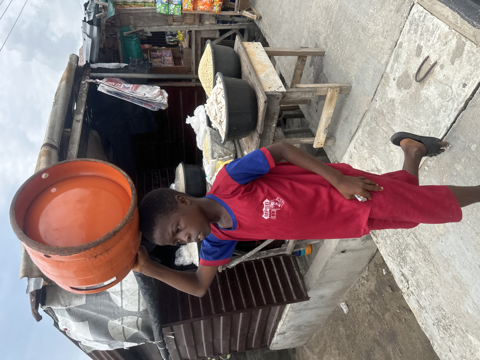 This image captures a young male carrying an orange liquefied petroleum gas (LPG) cylinder on his head, balanced with one hand, in what appears to be a market street setting in Badagry, Nigeria.
The subject is a young person with dark skin and short dark hair, wearing a red t-shirt with blue sleeves and collar, and red shorts. On the left chest of the t-shirt, a logo is visible, which includes the letters "MPSB" and a graphic of a building with books, suggesting a school or organization. He wears a single black slide sandal on his right foot, while his left foot is bare. His gaze is directed downwards as he balances the heavy cylinder.
The scene is an outdoor market stall or small shop. Behind the person, a rustic shop structure is visible, featuring corrugated metal roofing, wooden supports, and shelves. Inside the shop, several items are displayed for sale: two large black basins contain different foodstuffs, one filled with white items resembling processed cassava or garri, and another with yellowish grains or beans. Various packaged goods, including snacks and other provisions, are stacked on shelves. Wooden stools or makeshift tables are also present. The ground is a concrete pavement, showing cracks and some wear, with a metal staple-like object embedded in it. The sky is overcast, indicating a cloudy daytime setting. Faint text "1107 H" is visible on a concrete surface near the bottom right.