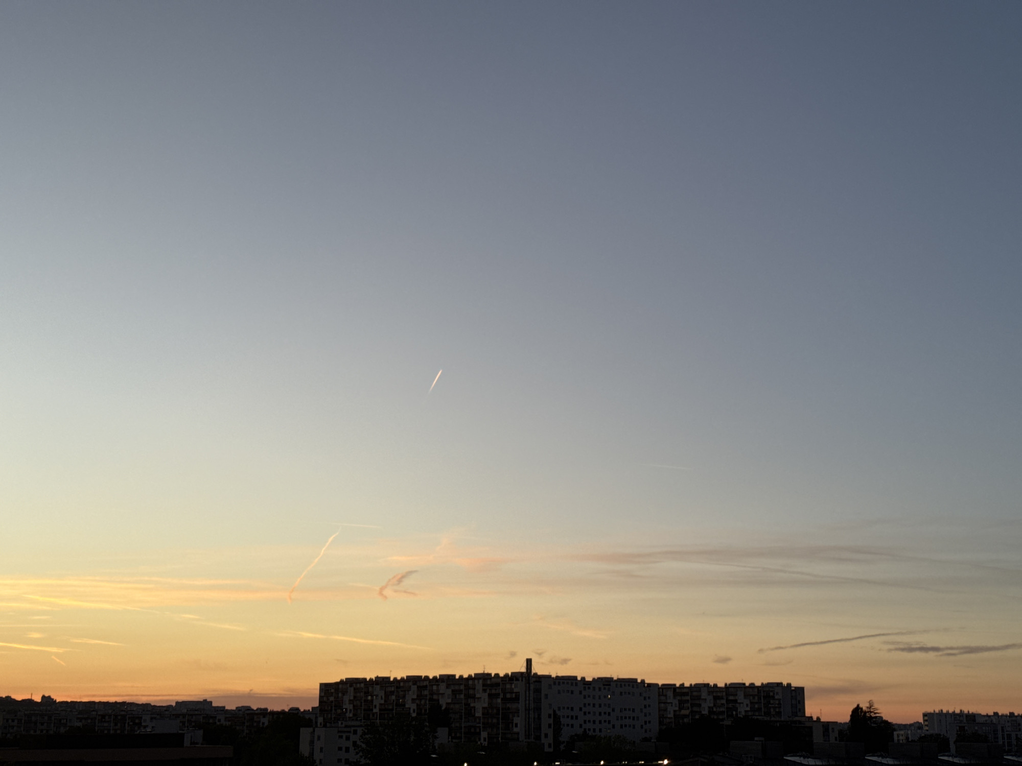 The image captures a twilight sky above an urban skyline. The sky transitions from a pale blue at the top to hues of orange and yellow near the horizon, indicative of sunset. Several contrails from aircraft are visible in the sky. Silhouetted against the colorful sky are rows of apartment buildings and other structures that form the city's architecture. The lighting suggests it is either dawn or dusk. The scene is devoid of any people or active interactions.