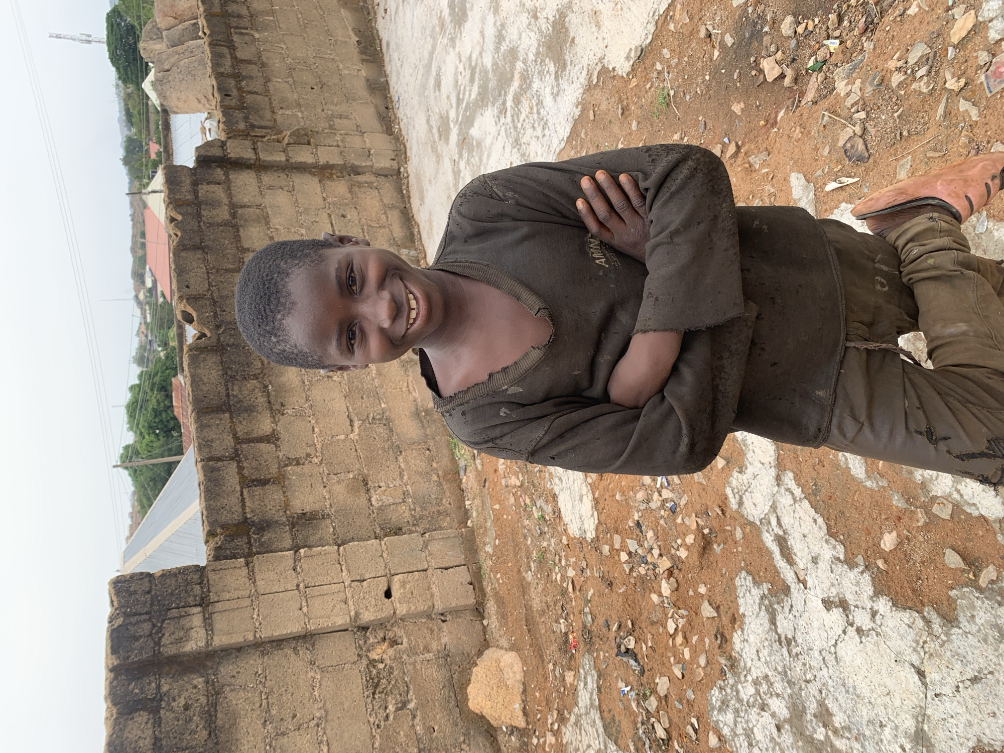 A young person is standing outdoors near a stone wall and a partially paved ground surface with dirt. The person is wearing dark, soiled clothing and leather shoes, with arms crossed, and smiling. The location is Gura Top, Nigeria. Other buildings and green trees are in the background.