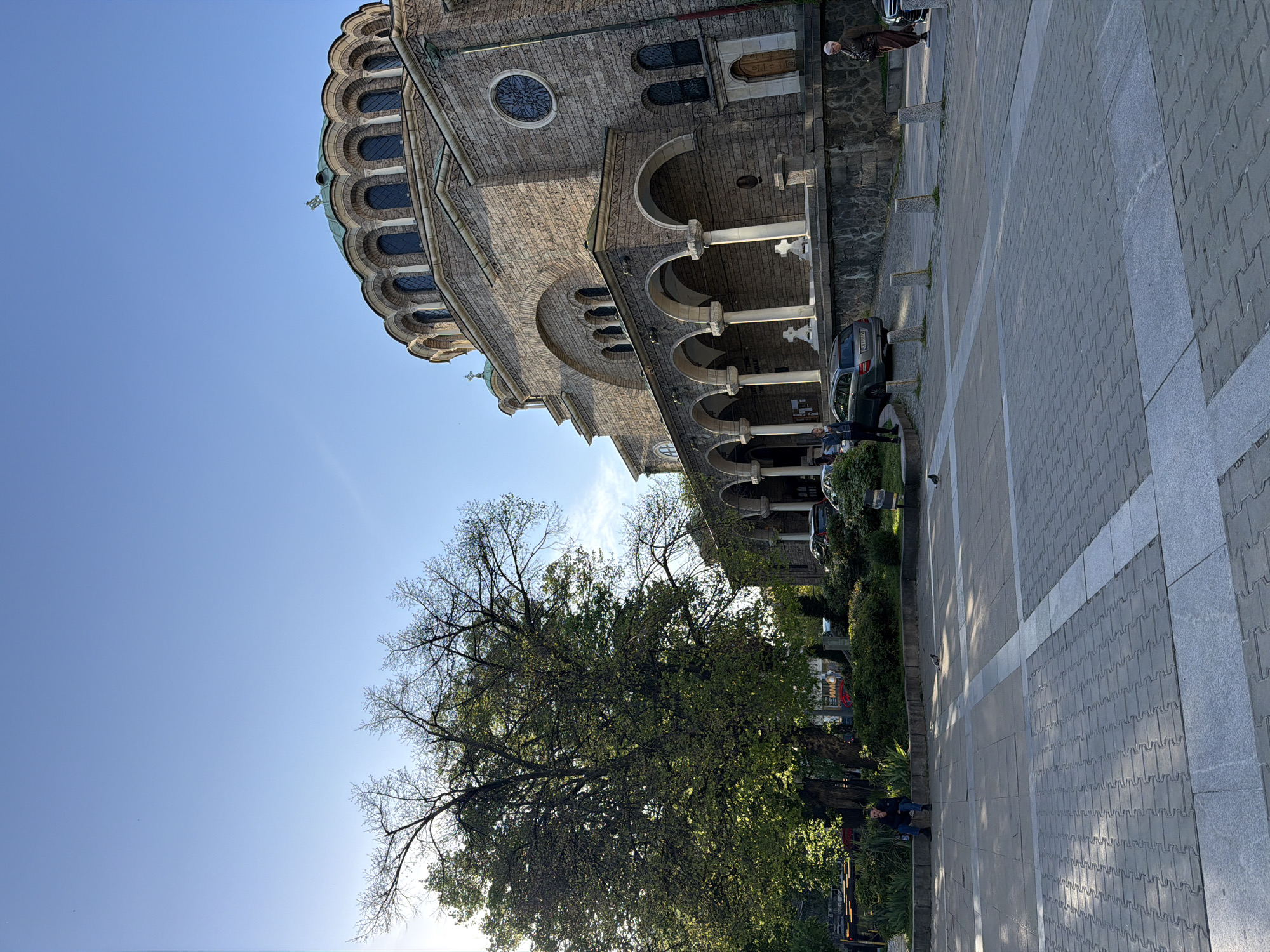 An outdoor wide shot captures a grand stone church, a paved plaza, and a large tree under a clear blue sky in Sofia, Bulgaria.
The prominent subject is a multi-domed building, likely an Eastern Orthodox church or cathedral, constructed from light-colored stone. Its facade features numerous arched windows, a large circular stained-glass window, and a portico with white columns supporting decorative arches. Green-domed roofs, topped with crosses, are visible against the sky.
In front of the church, a wide plaza is paved with grey interlocking pavers, interspersed with lighter grey stripes creating pathways. Small, light-colored stone bollards or low pillars line the edge of the plaza near the building.
A large, mature deciduous tree dominates the left side of the frame, displaying a mix of green foliage and bare branches. Several people are present: one individual in light clothing and a head covering stands near the church's entrance, while another sits on a low wall or bench by the tree. Other figures are indistinctly visible in the background. A silver or grey sedan is partially parked near the church, with a Bulgarian license plate (starting with "CA") faintly visible.
The scene is bathed in bright sunlight, casting distinct shadows, indicating a clear, sunny day, likely during midday or early afternoon. The overall impression is one of a calm, public urban space.