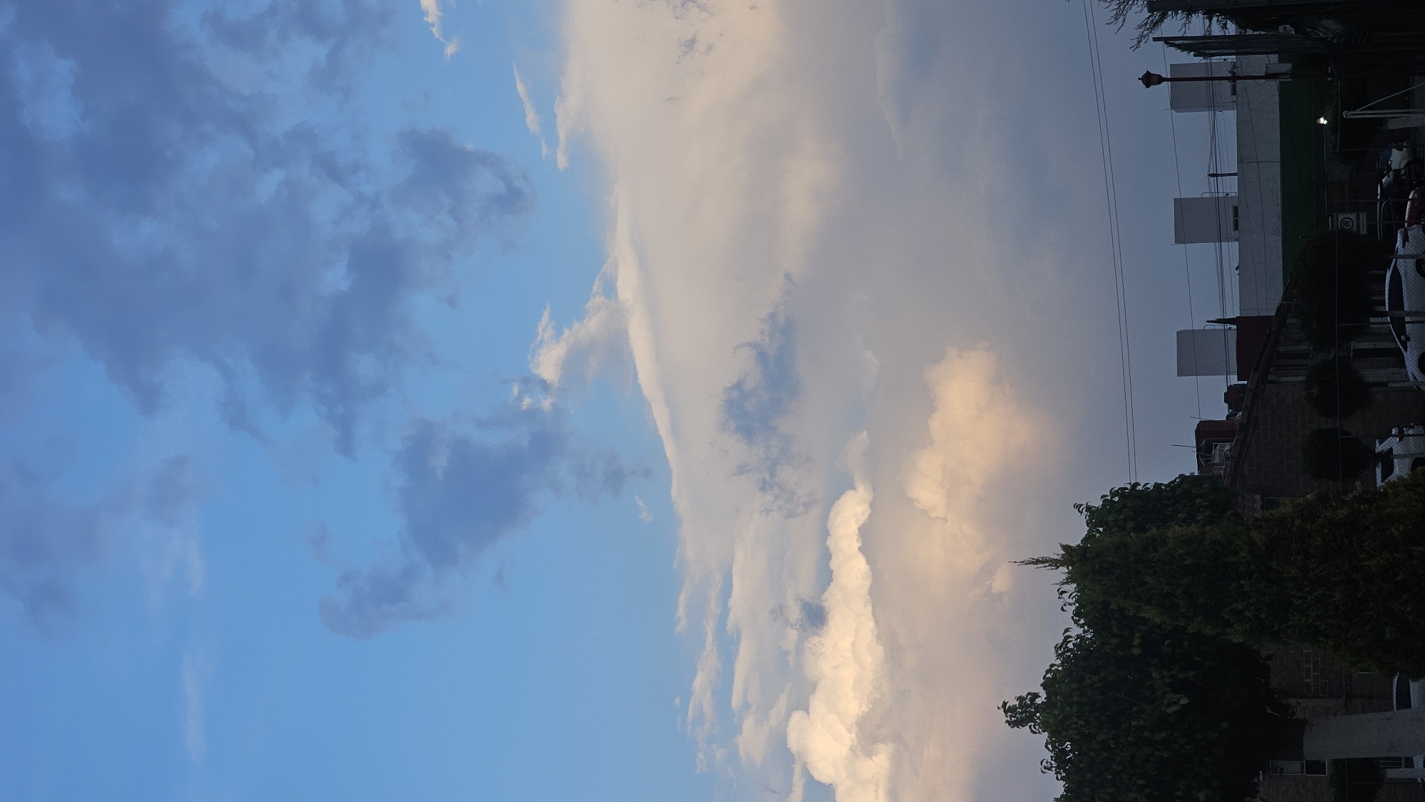 The image shows a sky with varying cloud formations, ranging from dark gray to light, almost white clouds, against a blue sky. The lower right portion of the image includes greenery, appearing to be trees and shrubs. In the background, there are buildings. There are power lines visible against the sky, suggesting this scene is in a populated or urban area. Based on the location information provided (San Bernardino Tlaxcalancingo, Mexico), the buildings in the background likely represent local architecture.