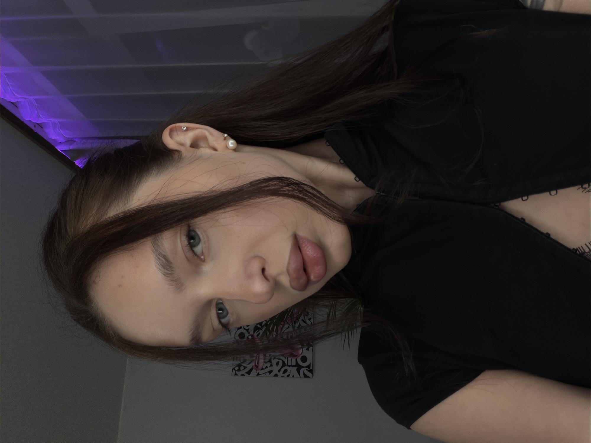 A woman with long brown hair, wearing a black top, is centered in the frame. She has fair skin, blue-grey eyes, and full lips. Her head is tilted to the side, and she is looking directly at the camera. She has multiple piercings on her left ear. In the background, there is a wall and a small piece of art, along with a purple-lit window. The specific location in Moscow, Russia, is not identifiable from the image.