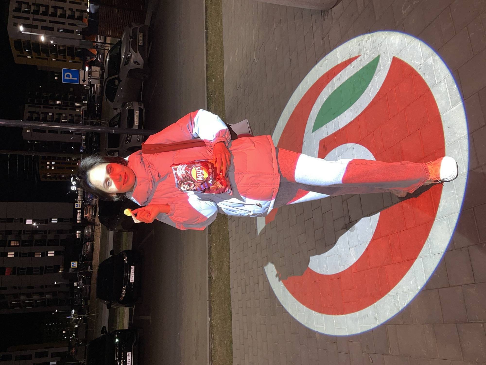 A woman stands on a paved surface, illuminated by a projected image of a red and white logo. She holds a bag of Lay's potato chips in one hand and a single chip in the other. She is wearing a red jacket, light-colored pants, and orange shoes. The logo is circular, featuring a red outline, a white interior, and a green leaf-like shape.
In the background, there are parked cars, streetlight, and a tall apartment building with illuminated windows. The scene is set at night. The location is most likely in the city of Pervomaysky, Russia, given the Cyrillic text on the chip bag.