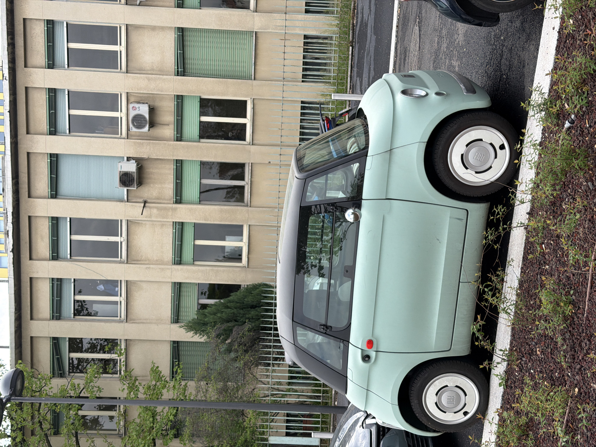The image, rotated 90 degrees counter-clockwise, features a light mint green/teal Fiat 500e electric car parked parallel to a sidewalk in an urban setting. The car occupies the right side of the frame, with its front and side clearly visible. Its silver wheel caps prominently display the "FIAT" logo.
Behind the car and along the left side of the frame, a multi-story building with a beige, textured concrete facade extends horizontally. The building features multiple rows of identical rectangular windows, many equipped with external rolling shutters in shades of light green and blue-grey, some partially open, others closed. Two white outdoor air conditioning units, one branded "LG," are mounted on the building's exterior. A white metal fence with pointed tops is visible between the building and the street.
In the foreground, to the right of the car, is a narrow strip of land covered with dark brown mulch and sparse green leafy plants, separated from the asphalt street by a white curb. A dark-colored vehicle is partially visible parked behind the mint Fiat. In the bottom left, a black streetlamp and some green trees are visible.
The scene depicts a daytime environment, with soft, diffused lighting suggesting an overcast sky. The overall setting is an urban streetscape, likely in Milan, Italy. No people are visible.