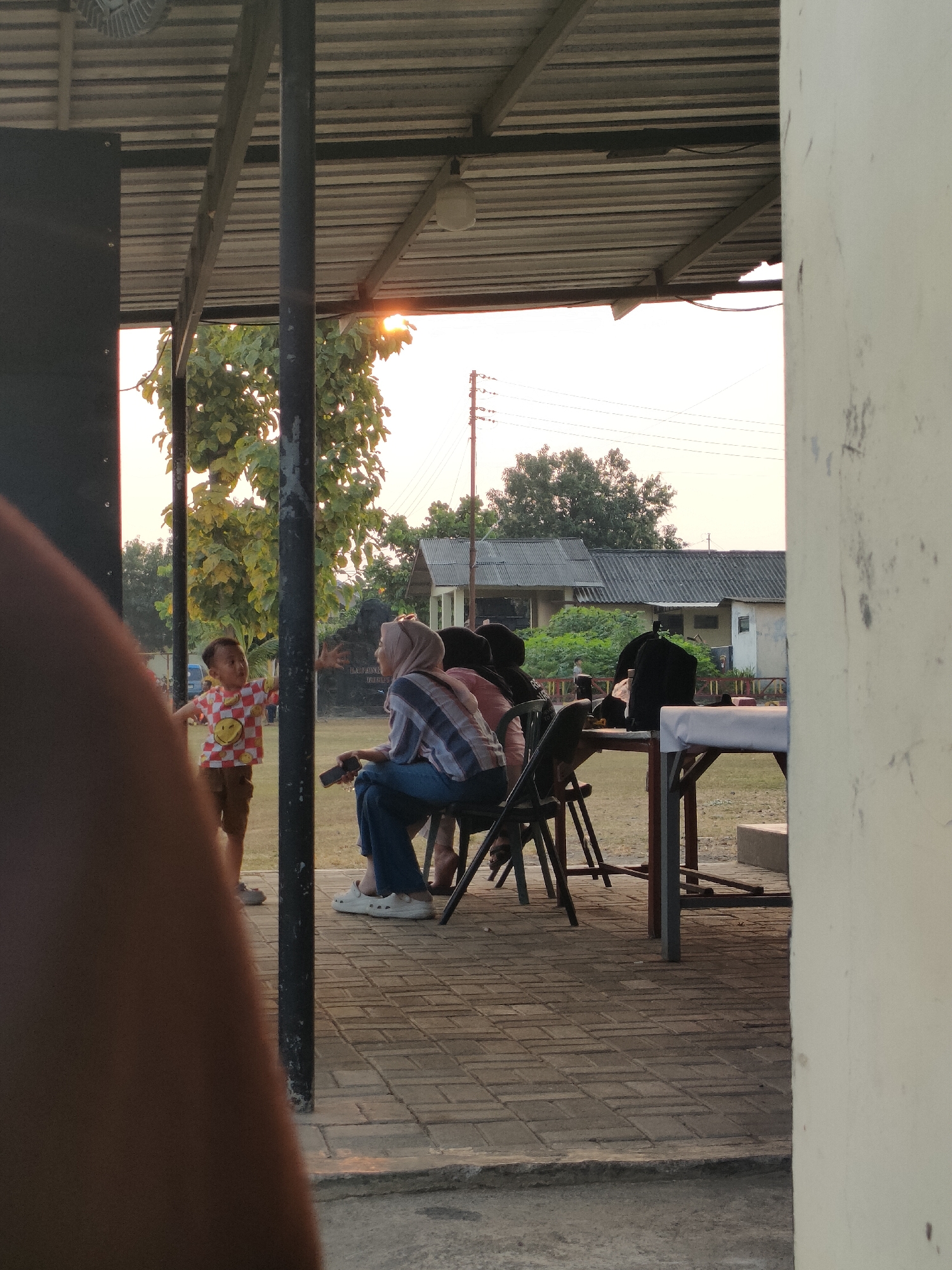 The image depicts an outdoor scene in Banguntapan, Indonesia, likely in a community gathering area or a park. The setting features a covered outdoor space with a metal roof supported by dark poles. In the foreground, a person's arm and shoulder are blurred, partially obscuring the view.
Several individuals are present. A young child, wearing a checkered shirt with smiley faces and brown shorts, stands with arms outstretched. To the right, a woman wearing a hijab and a plaid shirt sits on a chair, holding a smartphone. Other individuals are seated at a table covered with a white cloth, with their backs to the camera. Some are wearing dark head coverings.
The ground is paved with brick-like tiles. In the background, trees with green leaves are visible, along with buildings that appear to be houses or low-rise structures with grey tiled roofs. Utility poles and electrical wires stretch across the sky. The lighting suggests it is late afternoon or early evening, with the sun casting a warm glow. The overall atmosphere appears relaxed and informal, possibly a social gathering or a break time activity.