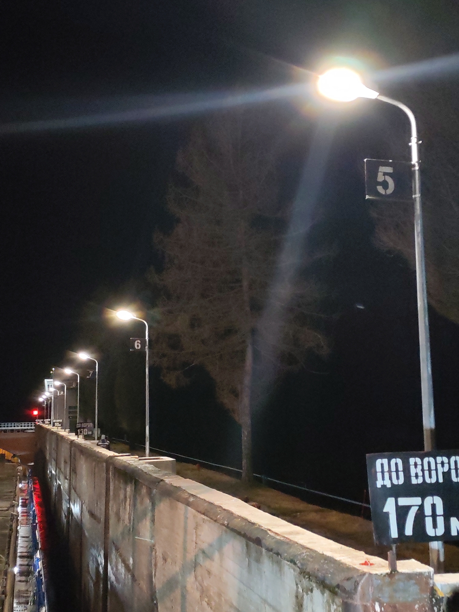 The image shows a night scene in an unidentified city. A concrete structure, possibly a dam or a canal wall, runs along the lower portion of the frame. Along the top of the structure, a series of evenly spaced streetlights illuminate the area. Each streetlight is numbered: visible numbers include 5, 6, and 7. A black sign reads "ДО ВОРОТ 170 M." A tree stands in the background between streetlights, partially silhouetted against the dark sky. Red lights are visible on the left side of the frame, possibly indicating traffic control or safety signals on the dam. The environment appears dark and calm.
