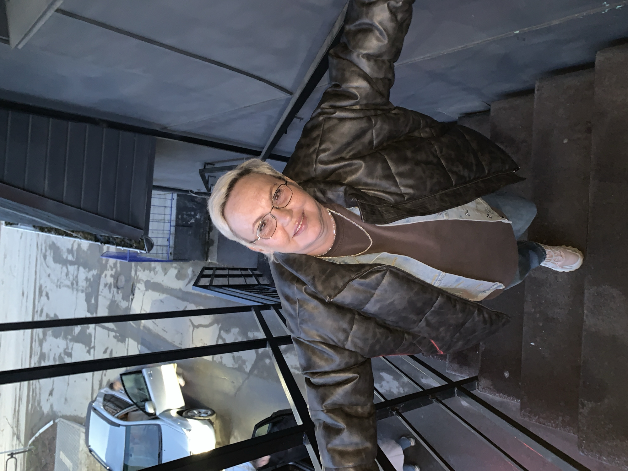 The image captures a person standing on a dark, multi-level outdoor staircase, smiling up towards the camera. The individual is an older person with short, light-blonde hair and glasses, wearing a dark brown puffer jacket, a brown t-shirt, blue jeans, and light-colored sneakers. A silver chain is visible around their neck.
The setting appears to be an outdoor stairwell or passage in an urban environment. The stairs are dark, possibly concrete or stone, and feature black metal railings. Below the stairs, an outdoor area is visible with what appears to be wet or damp concrete pavement, suggesting recent rain or moisture. A silver compact car is parked on this lower level, with its driver's side door open. The surrounding structures have a utilitarian, possibly industrial, aesthetic with grey building materials.
The time of day appears to be daytime, though the lighting is somewhat muted, possibly due to an overcast sky or the shadows of surrounding buildings. The person's smiling expression conveys a positive mood. No visible text is present in the image. The location is Chaikovsky, Russia.