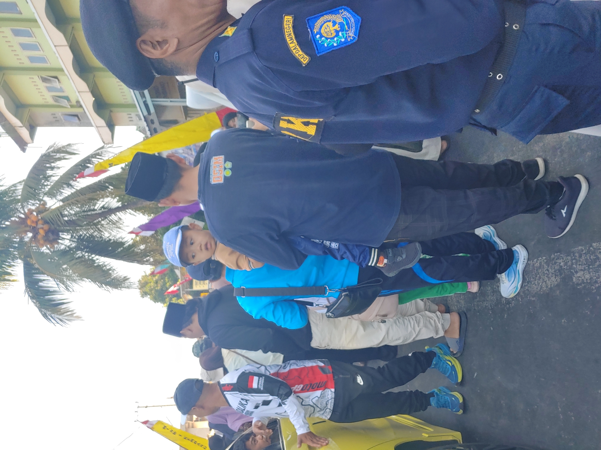 The image shows a group of people standing on a street, likely in Kembang Kerang, Indonesia, as indicated by the provided context. The scene appears to be a public gathering or event. In the foreground, a person in a dark blue uniform with patches is facing away from the camera, partially obscuring the view of the people behind them. The uniform has a patch on the sleeve with text that appears to say "DEP. DALAMNEGERI."
To the left of the uniformed individual, a child is being held by another person. The child is wearing a light brown shirt and a blue cap. Behind them, a crowd of people are visible, many of whom are wearing casual clothing. Some are wearing hats, including a black peci on one individual. There are also flags, one yellow and one purple, visible in the background, suggesting a festive or ceremonial occasion. A palm tree with coconuts is present in the upper portion of the image. The environment suggests an outdoor urban setting, with buildings visible in the background and a paved road surface. The overall lighting indicates it is daytime, possibly late afternoon given the quality of light.