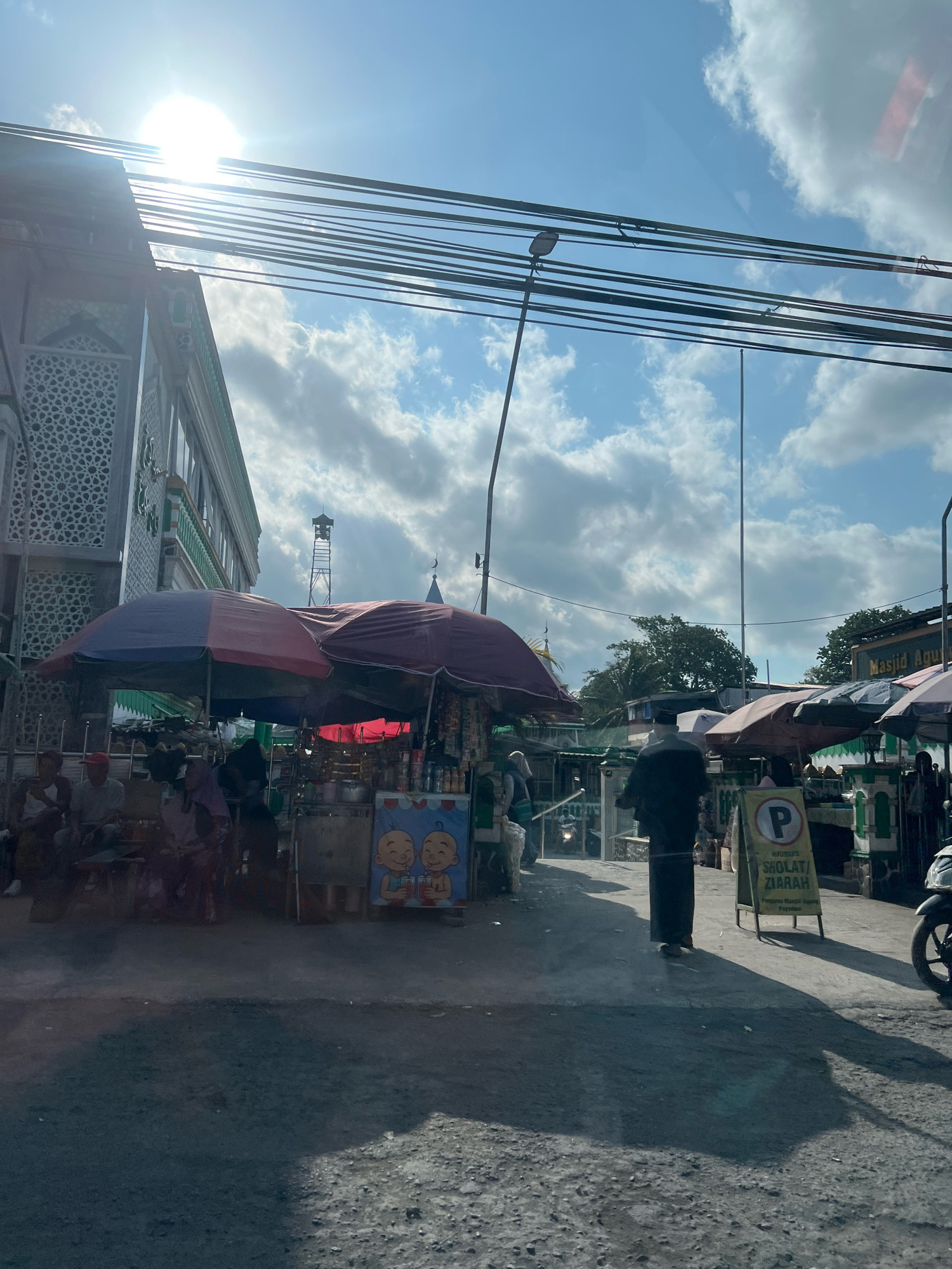 The image depicts a street scene in the city of Payaman, Indonesia, on a bright, sunny day with scattered clouds. In the foreground, a paved road leads towards a row of shops and market stalls. Several people are visible, including individuals sitting under umbrellas and a person walking away from the camera dressed in traditional Islamic attire. Market stalls display various goods, with colorful umbrellas providing shade. A sign with a "P" and text in Indonesian, likely indicating parking or a specific area, is placed near one of the stalls. In the background, a building with intricate geometric patterns and a green trim is visible, suggesting it might be a mosque or a significant cultural building. The overall atmosphere appears to be that of a bustling local market or a common area outside a religious site, with everyday activities taking place.