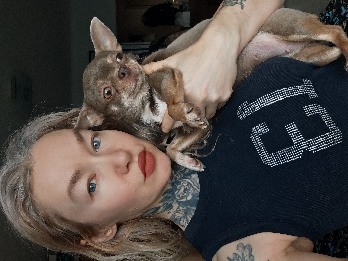 The image displays a close-up indoor portrait of a female individual holding a small dog. The female has fair skin, light blue eyes, and light blonde hair with visible grey roots, and wears reddish-brown lipstick. She exhibits extensive tattoos, including a floral pattern covering the left side of her neck and upper shoulder, and a partial design visible on her left upper arm. She is dressed in a dark, ribbed sleeveless top featuring the letters "EJ" rendered in silver rhinestones on the chest. She holds a small, short-haired dog, resembling a Chihuahua, with light brown fur and lighter markings on its chest and paws. The dog possesses large, erect ears and dark eyes; its visible underside displays four distinct nipples. Both subjects are looking directly towards the viewer. The background is dark, out of focus, and non-descriptive, consistent with an interior setting. There are no visual elements within the image that allow for the determination of its location in the city of Saint Petersburg, Russia, or any other specific geographic area.