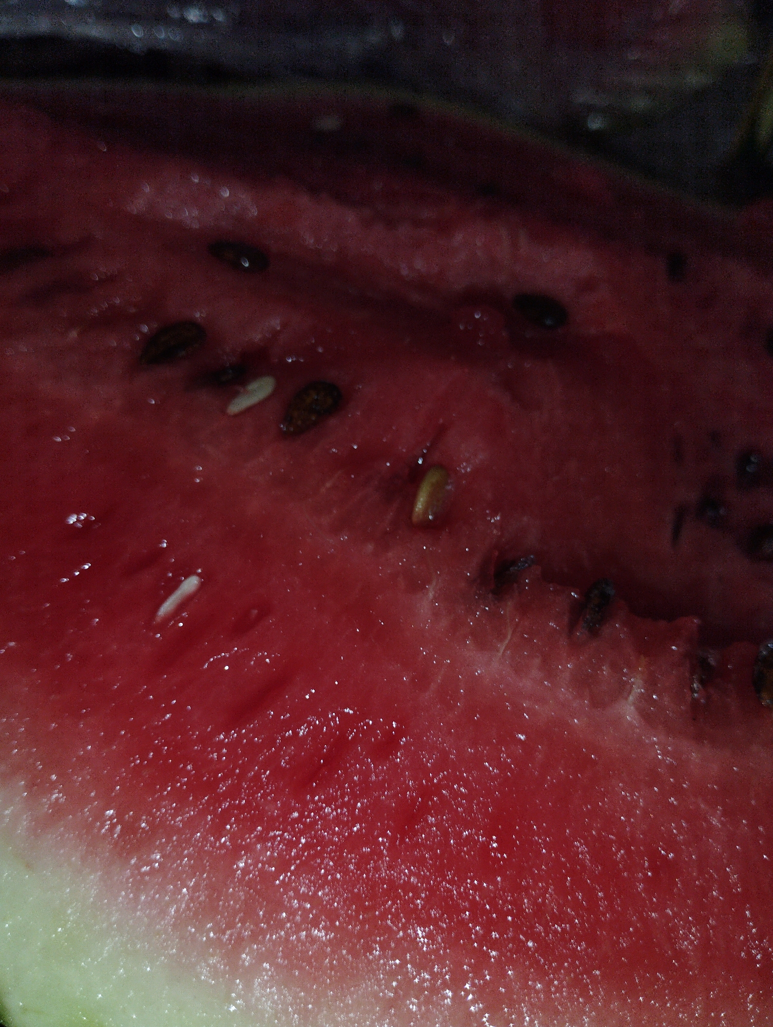 A close-up, high-angle shot shows the glistening, red flesh of a sliced watermelon, revealing numerous black and some white seeds scattered throughout. The texture is moist and slightly fibrous, with highlights glinting off the surface. The rind, visible on the left, transitions from pale green to white. The background is dark and out of focus, suggesting an indoor setting or low light conditions. The image's context is not specified as being in the city of Гроздь, Kyrgyzstan, or any other specific location.