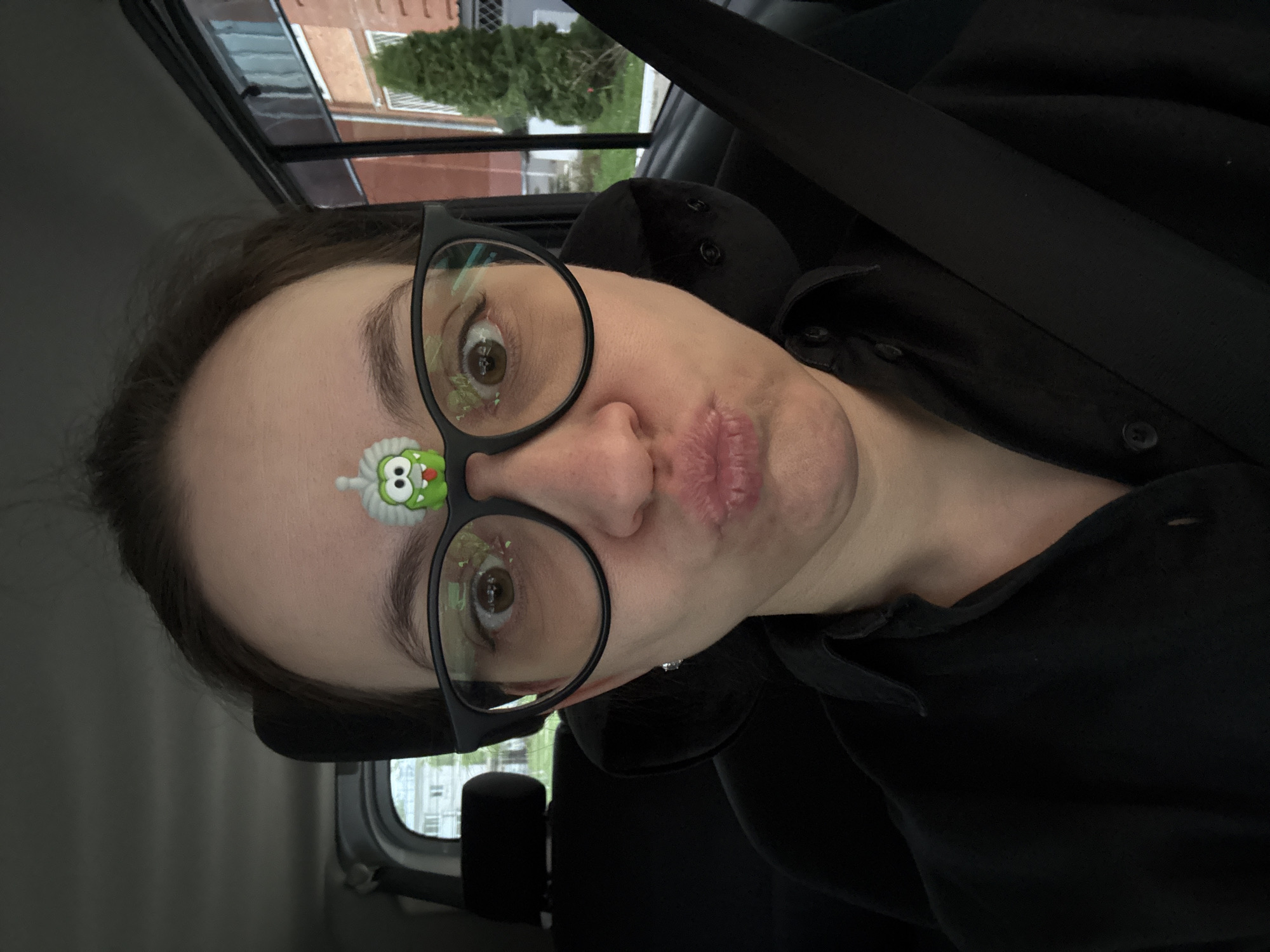 A woman is in a car wearing eyeglasses with a green sticker on her forehead. She is wearing a black shirt and a seatbelt. The exterior environment appears to be a residential or urban area, with a building visible in the window, partially obscuring the street outside. Due to the limited information, I cannot determine the location is in Chaikovsky, Russia.