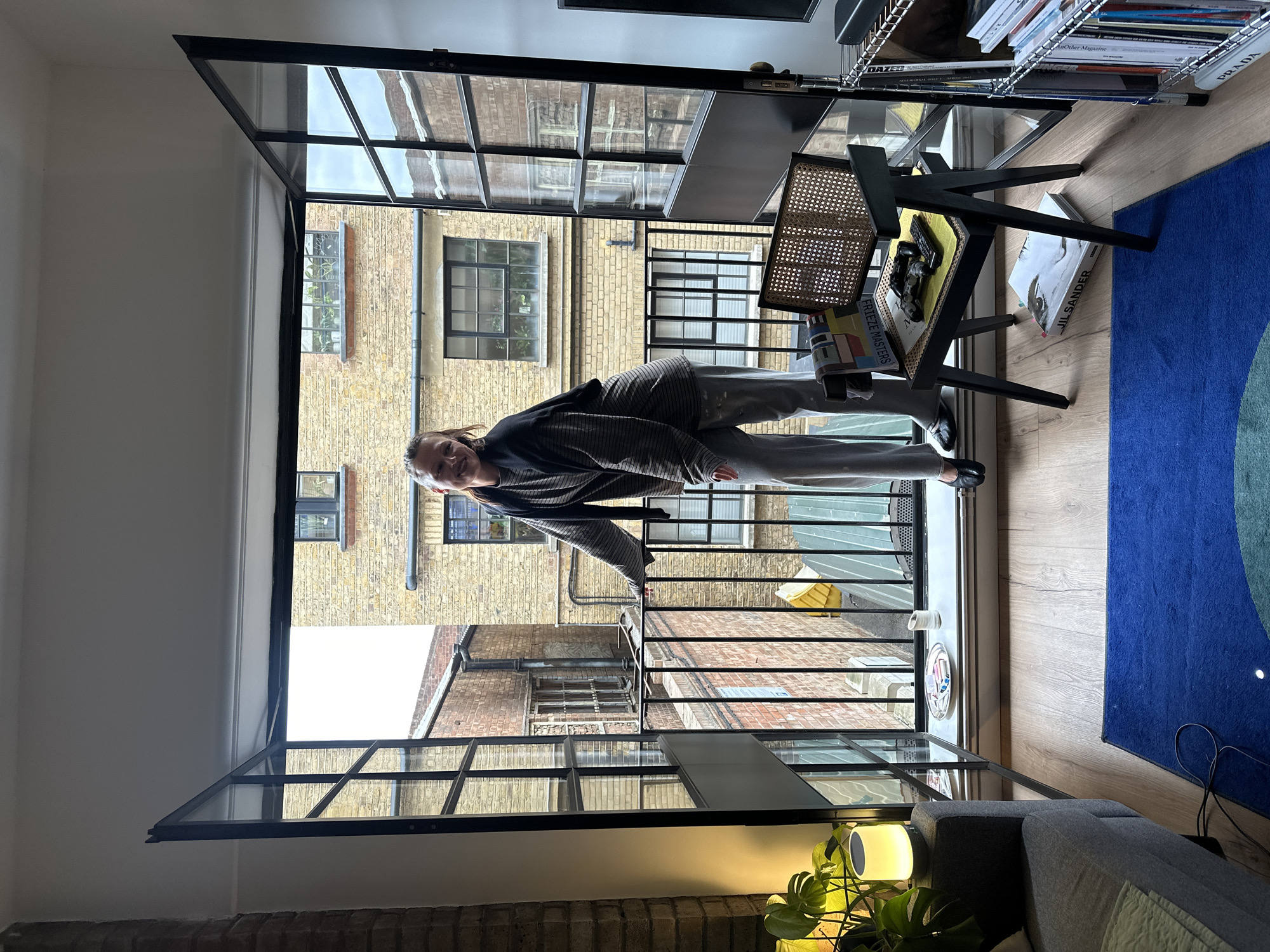 A person wearing a striped top and grey trousers stands on a balcony in a London flat. The balcony overlooks a brick building in what looks like a courtyard or narrow street. The flat features black framed windows and doors. Inside the flat, there is a desk with a wicker chair, a blue rug with concentric circles, and part of a grey sofa. Books are stacked on a shelf near the desk. A small lamp shines near the sofa.