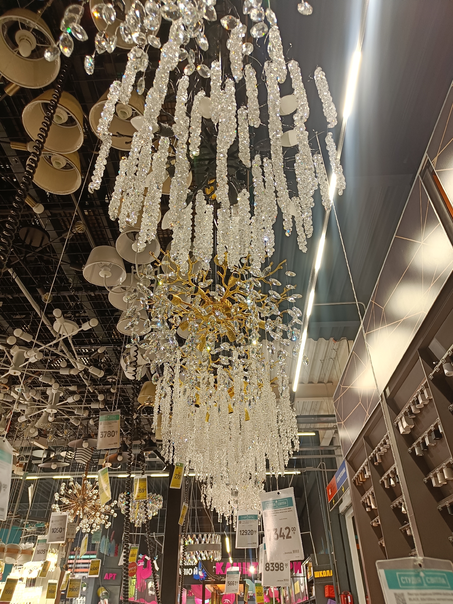 The image shows a display of chandeliers in a retail store, likely a lighting store or a department store with a lighting section. The main subject is a large, ornate chandelier with numerous clear, faceted crystals hanging from a golden frame. This chandelier is a focal point, with cascades of crystals creating a dazzling effect.
Surrounding the main chandelier are other lighting fixtures, including more chandeliers, pendant lights, and ceiling lamps. Many of these have price tags attached, suggesting a sale or retail environment. Some of the visible text on the price tags includes prices in local currency, such as "3780.00", "12920.00", "342.00", and "8398.00". There are also labels with product names or codes, like "APT" and "ШКОЛА", which might indicate product categories or brands.
The setting appears to be indoors, under artificial lighting. The ceiling is a dark color, possibly black or dark gray, with a grid-like structure visible in some areas. The overall atmosphere is one of a well-lit commercial space showcasing a variety of decorative lighting options. The image is a close-up, focusing primarily on the chandeliers and their intricate details, with the background elements slightly blurred. No people are visible in the frame. The time of day and weather are not discernible from the image. The location context provided is Vinnytsia, Ukraine.
