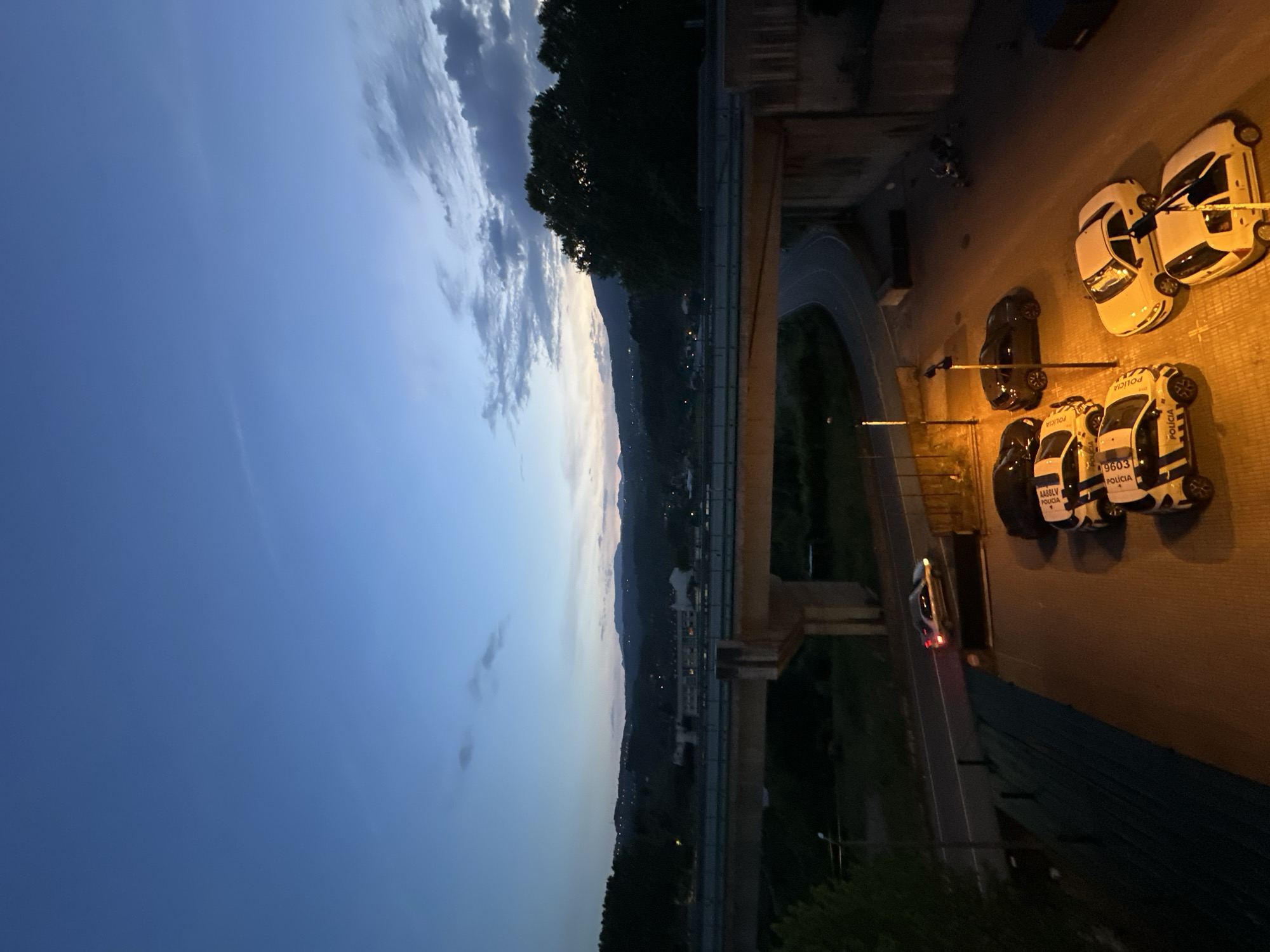 An elevated, low-angle, dusk shot depicts a bridge and a parking area with several parked vehicles. The sky transitions from a deep blue in the upper left to a lighter hue near the horizon, with scattered clouds. A dark treeline extends along the horizon. The bridge is visible at the bottom of the frame, spanning a valley where lights from a town or city are discernible.
To the right of the bridge, a parking area houses several vehicles. These include what appear to be police cars (indicated by the word "POLÍCIA" written on the side) and standard civilian vehicles. One vehicle has its headlights illuminated.
Based on the presence of the word "POLÍCIA" in Portuguese and further markings on the vehicles, it is likely the location is Vila Real, Portugal.