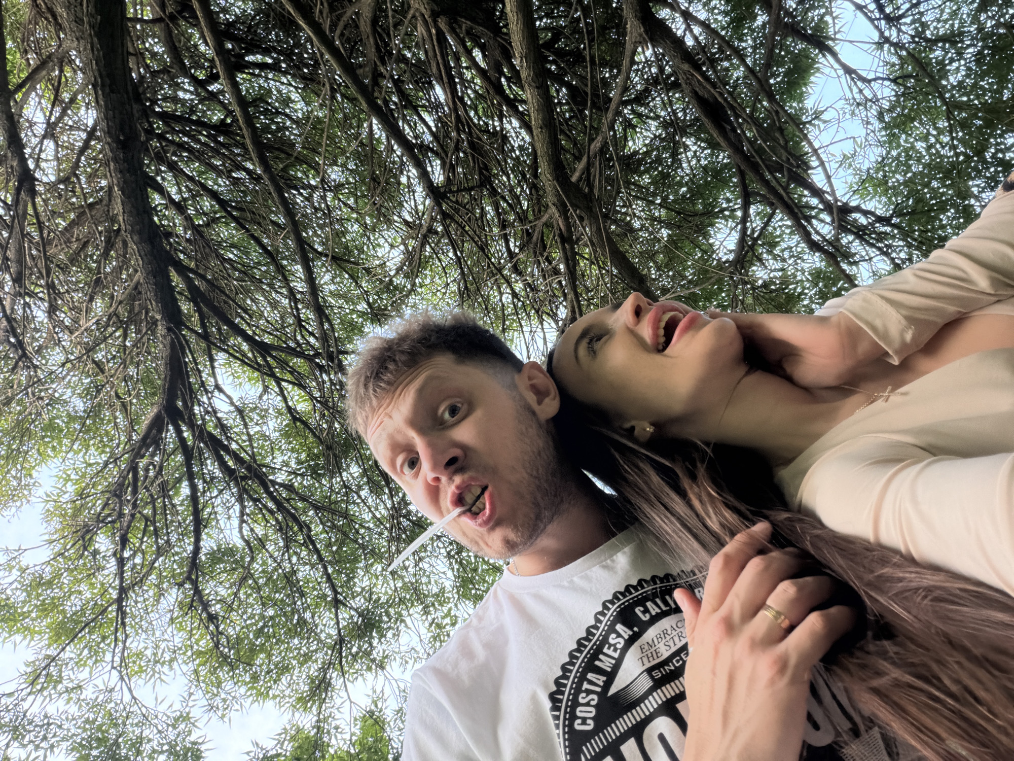 An outdoor, low-angle shot captures two individuals from below, looking up through a canopy of tree branches and green leaves against an overcast sky in Unknown, Russia.
The foreground features a light-skinned male on the left, wearing a white t-shirt with a black and white "COSTA MESA, CA" graphic. He has short light-brown hair and stubble, with wide-open eyes gazing directly at the camera. A clear, tubular object is horizontally positioned between his teeth, protruding from the left side of his mouth. His right hand, adorned with a gold band on the ring finger, rests on the upper back or hair of the second individual.
To the male's right and slightly below is a light-skinned female with long brown hair, wearing a light-colored, long-sleeved top and a gold cross pendant necklace. A small gold stud earring is visible in her right ear. Her head is tilted upwards and to the right, and her mouth is open, conveying an expression of mirth. Her right hand is raised, with fingers gently touching her jawline.
The background consists of a dense network of dark, textured tree branches and scattered green foliage, with patches of bright, diffused sky visible between the leaves.
