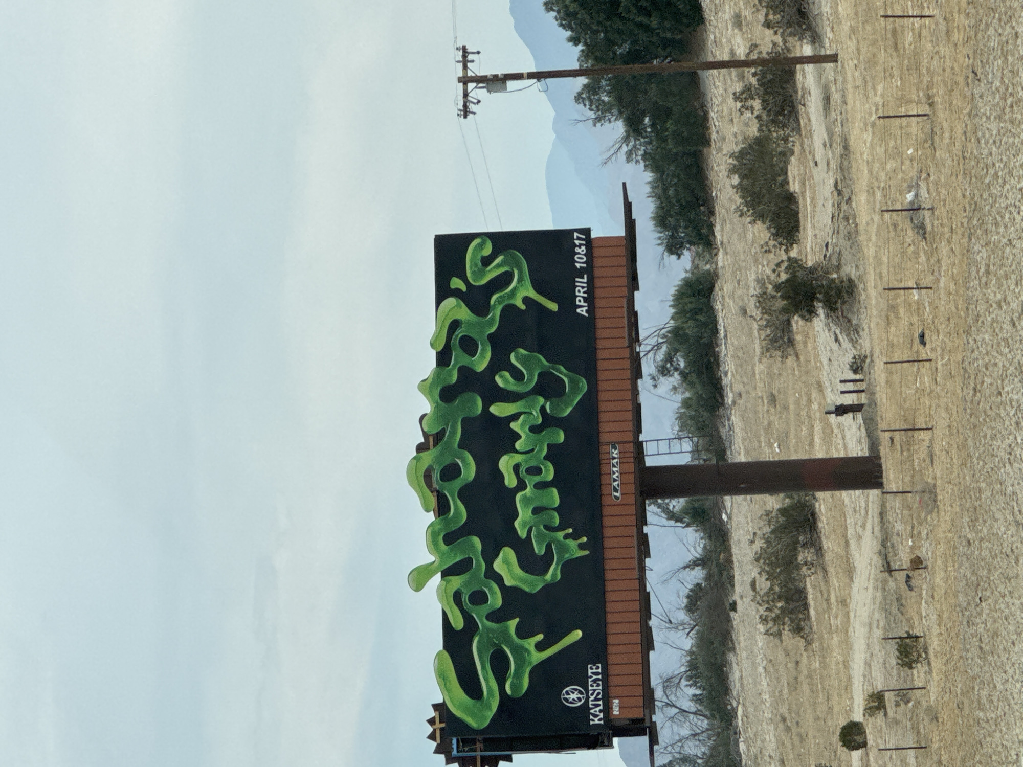 A billboard in Cathedral City, United States of America displays green, blob-like text against a black background, advertising dates for April 10th and 17th. The billboard is sponsored by Katseye and Lamar. The surrounding environment is arid with sparse vegetation, set against a backdrop of mountains and a hazy sky. A utility pole with power lines stands nearby. The billboard is oriented vertically in the image.