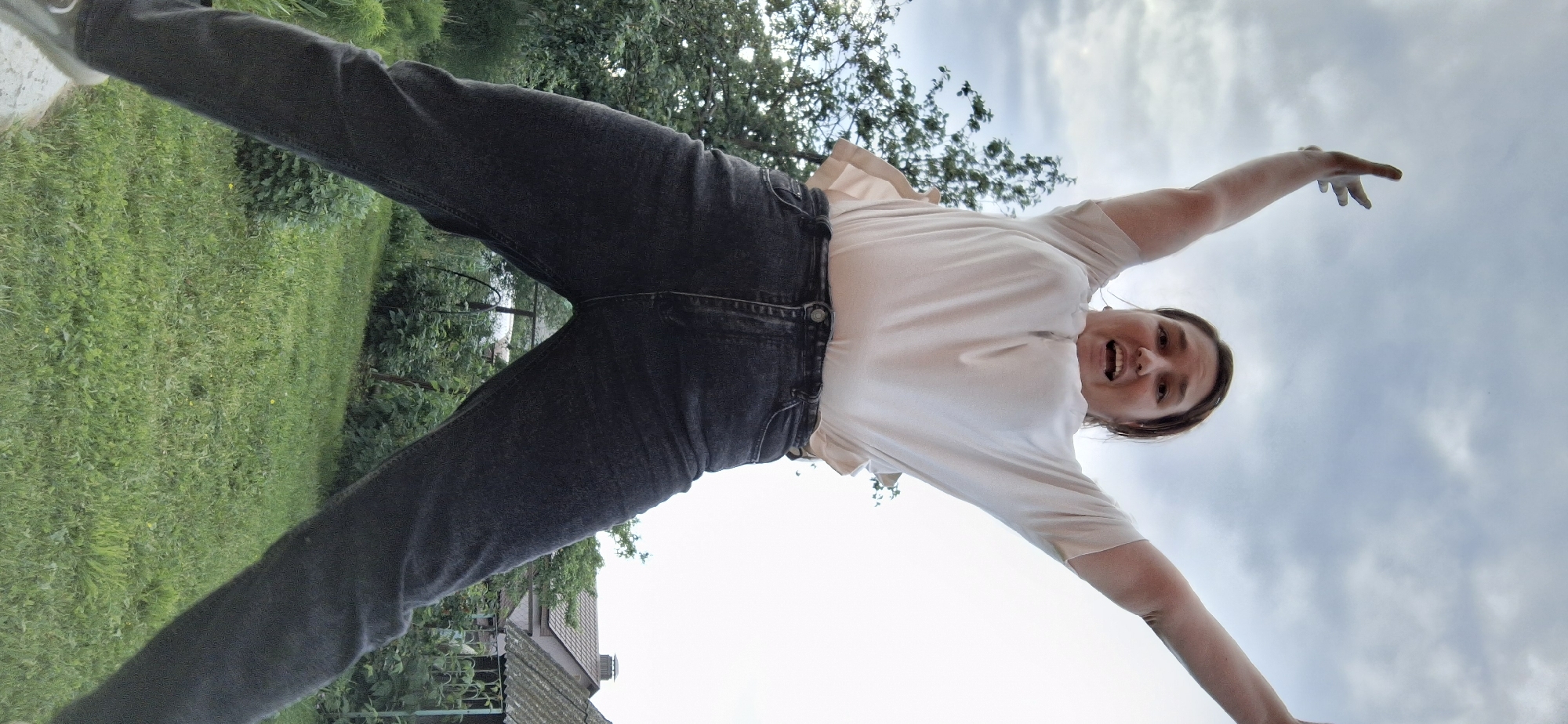 A medium shot captures a young woman in mid-air, jumping with her arms outstretched towards a cloudy sky. She is positioned in the foreground, her body angled slightly upwards, with her legs spread wide. The woman is wearing a light-colored short-sleeved shirt and dark denim jeans. Her facial expression is one of surprise or excitement, with her mouth open and eyes wide.
Behind her, a backdrop of lush green grass and dense foliage suggests an outdoor setting. Trees with green leaves are visible to the left, and a glimpse of houses with brown roofs and a fence can be seen in the distance on the right. The overall atmosphere is casual and energetic, suggesting a moment of playful activity. The location is identified as Chaikovsky, Russia.