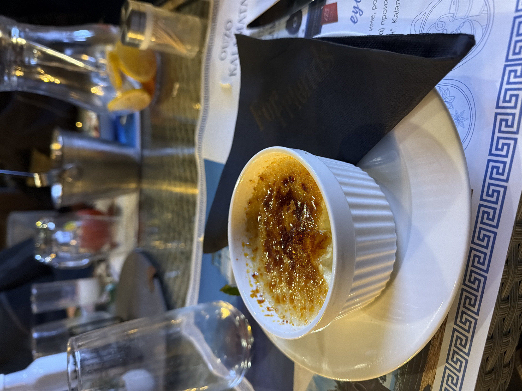 The image depicts a crème brûlée dessert served in a white ramekin on a white plate with a black napkin. The dessert is on a table at a restaurant, with visible glassware and other table settings. A placemat with Greek key design is under the plate. The location details, specifically Novakovtsi, Bulgaria, cannot be verified from the image. There is no specific information about people, actions or interactions that can be verified from the image.