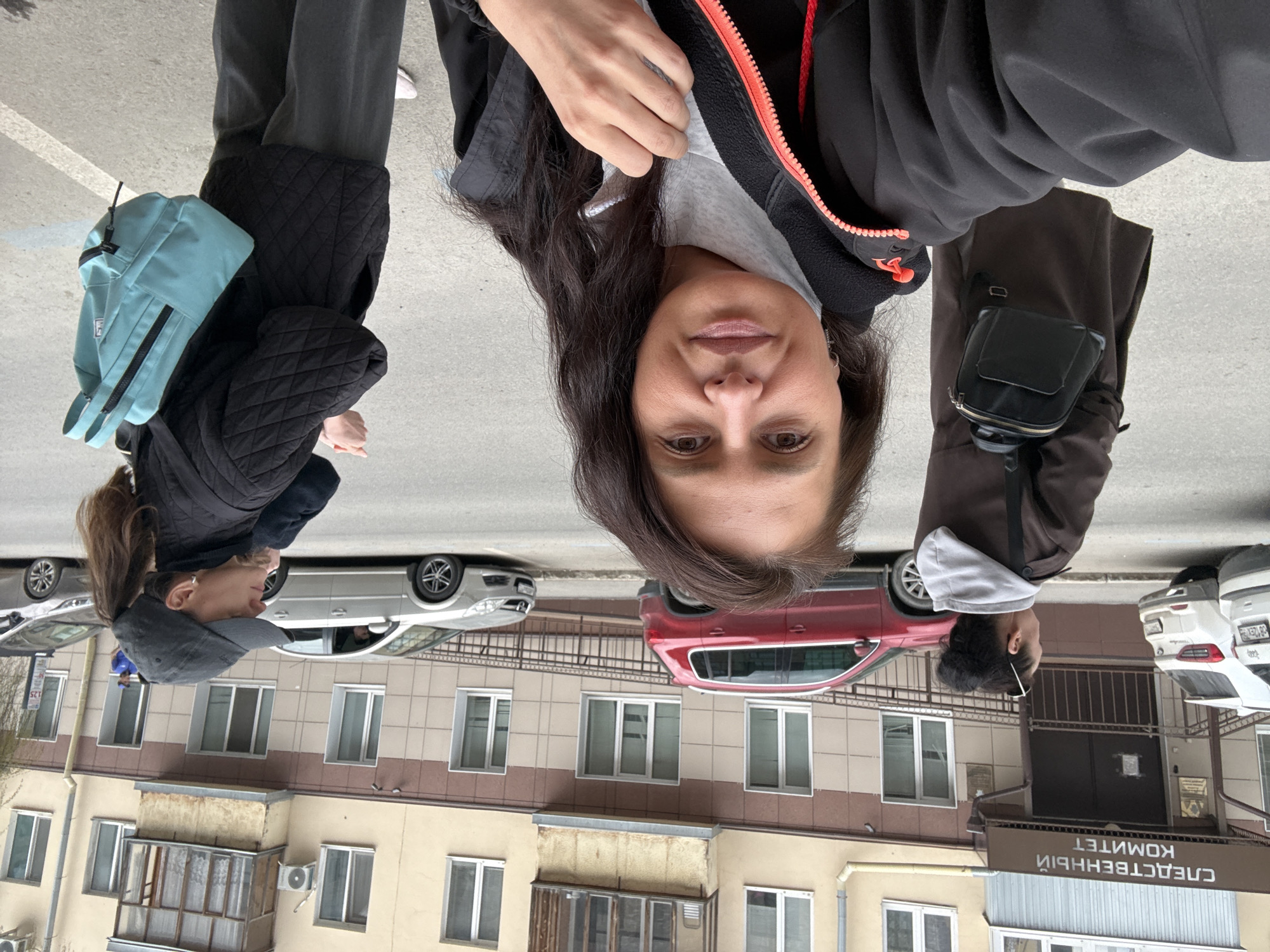 The provided image is rotated 90 degrees clockwise. When viewed upright, it captures an urban street scene in Kazan, Russia, on an overcast daytime.
In the immediate foreground, a woman is seen looking directly into the camera, appearing to take a selfie. She has long, dark, wavy hair and dark eyes, wearing a light grey t-shirt under a black jacket with a striking bright orange zipper. Her expression is neutral with a subtle hint of a smile.
To her left, another person is partially visible from the waist down, wearing dark trousers and a black quilted jacket, with a light blue backpack resting on their shoulder. On her right, a third individual is visible, wearing a brown jacket and carrying a small black fanny pack or bag across their body. Further in the background on the left side of the street, a fourth person is briefly seen wearing a dark cap and top.
The background features a street lined with several parked cars, including a silver sedan, a red SUV, and two white vehicles further along. Behind the cars, a multi-story building with a light-colored facade, numerous windows, and several balconies rises. Air conditioning units are visible on the building's exterior. The ground is asphalt or concrete, marked with white parking lines.
A prominent detail on the building to the right is a brown sign with Cyrillic text. When viewed upright, it clearly reads "СЛЕДСТВЕННЫЙ КОМИТЕТ," which translates to "Investigative Committee." The overcast sky suggests cool weather, consistent with the jackets worn by the individuals. The scene portrays a casual moment in a city environment.