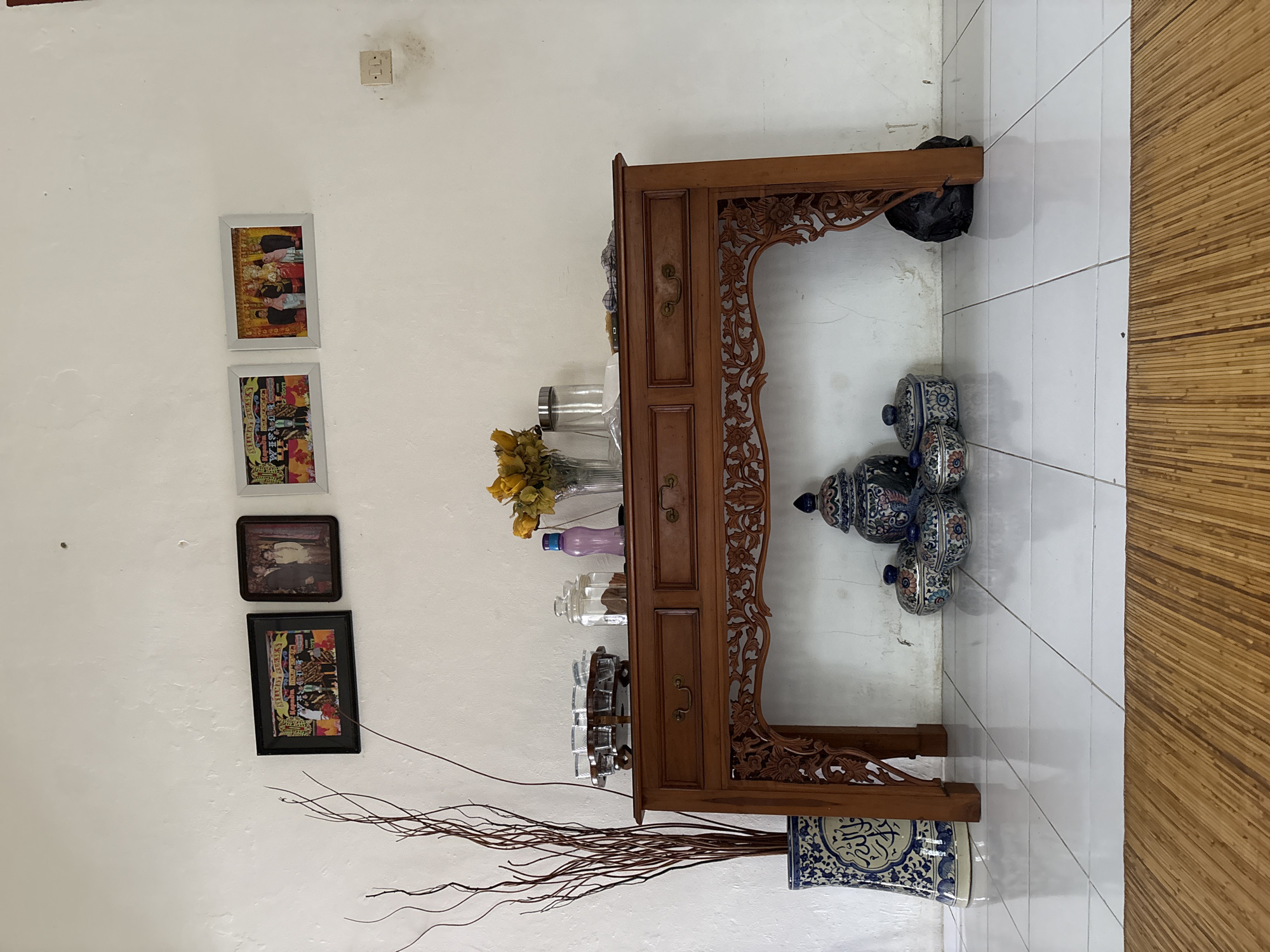 The image shows a console table against a white wall in a room, possibly in Uleepulo, Indonesia, given the stylistic elements of the decor. The table is made of dark wood and features three drawers with ornate metal pulls. The top of the table displays several items: a glass vase holding yellow flowers, a glass jar filled with dark-colored contents, a purple bottle, and a wooden tray holding multiple small glasses. To the left of the table, a tall blue and white patterned ceramic vase holds a bundle of dry, dark-colored branches. Below the table, a collection of decorative ceramic containers with lids, in blue and white patterns, are arranged on the floor. A large, dark blue and white patterned ceramic jar with a lid stands to the right of these smaller containers. To the far right, a black trash bag is partially visible behind the table leg. The wall above the table is adorned with three framed pictures. The floor is tiled with large white square tiles, and a woven straw mat is visible in the foreground. The lighting suggests an indoor environment with natural light coming from an unseen source.