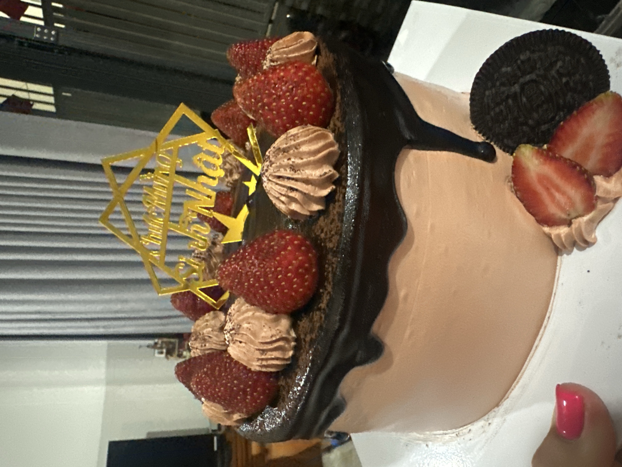 The image shows a cake decorated with strawberries, chocolate icing, and an Oreo cookie. A gold cake topper with Vietnamese writing is also present. The background is out of focus, but it appears to be an indoor setting, possibly a kitchen or dining area. A hand with red nail polish is visible in the bottom right corner. The location is stated as Bac Nha Trang Ward, Vietnam.