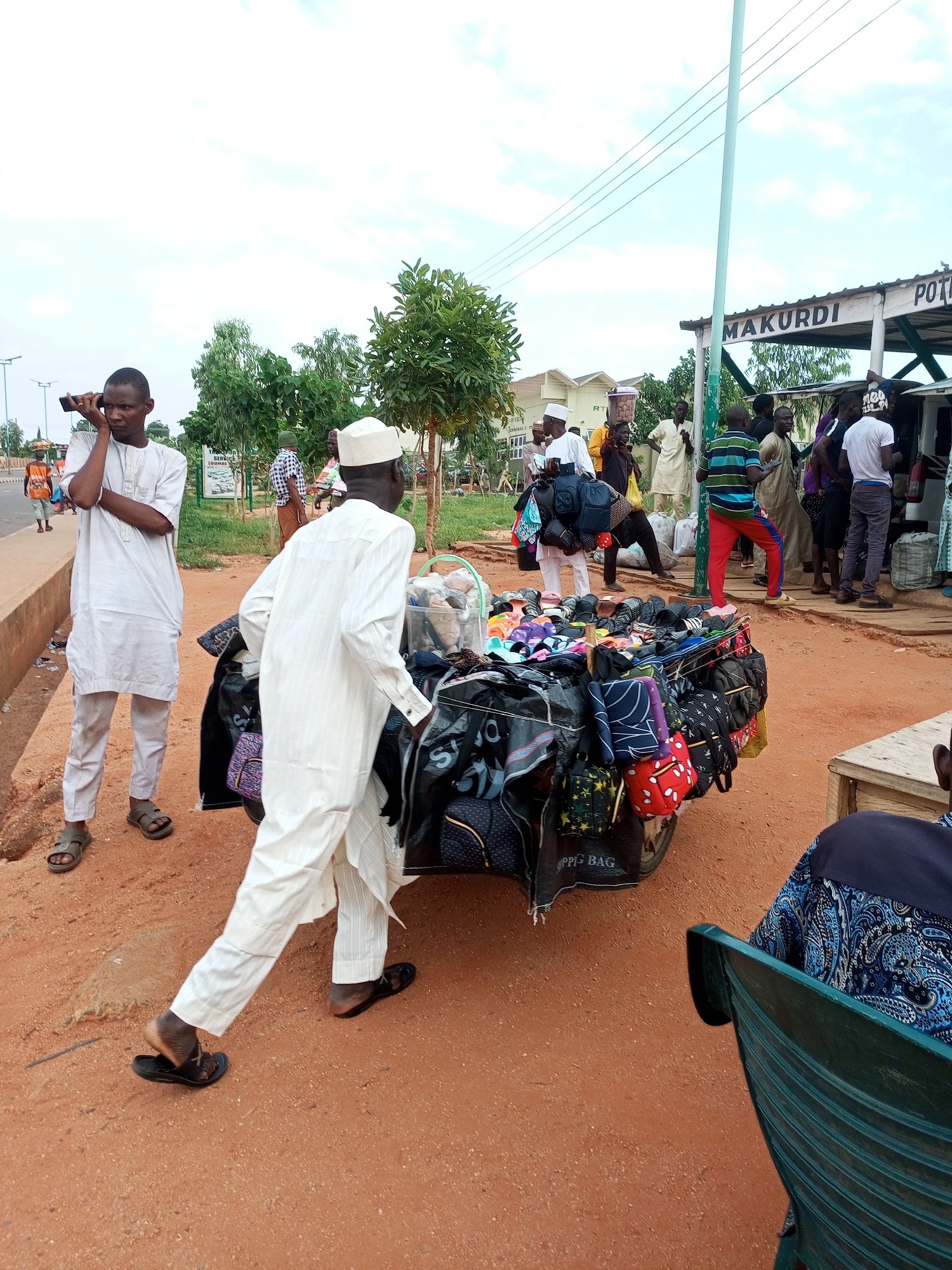 The image depicts an outdoor market scene likely in Gombe, Nigeria, near a bus park as suggested by the structure labeled "MAKURDI" which could indicate a destination or origin for transport. A man in white traditional attire is talking on a mobile phone, standing beside a road with a concrete barrier. Further into the scene, another man, also in white, walks past a cart laden with numerous bags and footwear, which appears to be a vendor's stall. A group of people are gathered near a vehicle and a covered structure, possibly a bus terminal. The ground is reddish earth, and sparse vegetation dots the background. The sky is overcast.