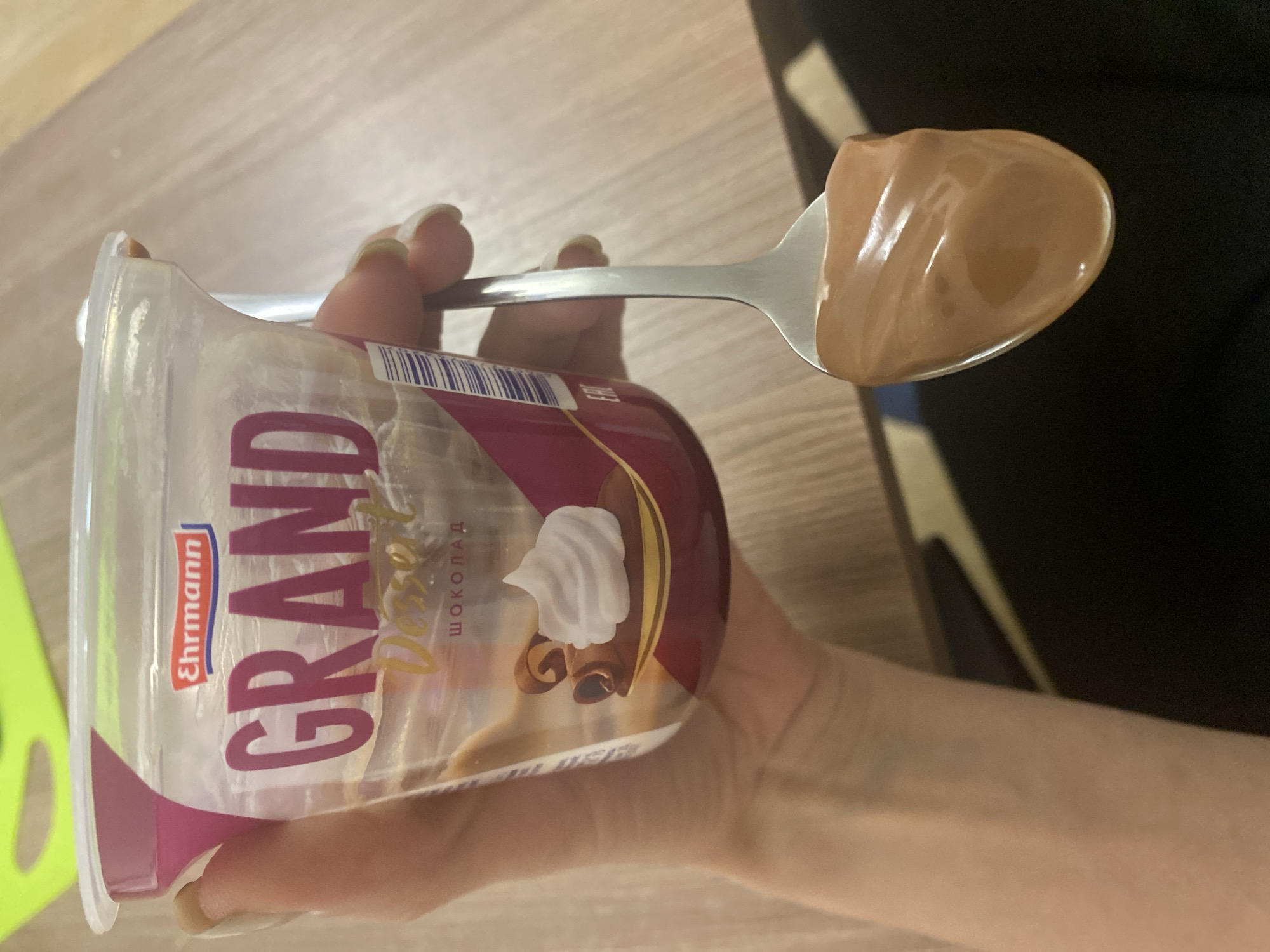 An overhead shot captures a person's hand holding a plastic cup of chocolate dessert and a spoon filled with the same creamy treat. The hand, with manicured fingernails, grips the clear plastic cup, which features a prominent label.
The label on the cup clearly displays the brand name "Ehrmann GRAND," with "Dessert" (partially visible) and "шоколад" (shokolad), meaning chocolate in Russian, below it. An illustration on the label depicts a dollop of white whipped cream and two chocolate rolls, suggesting the flavor or type of dessert. A barcode and the "EAC" mark (indicating conformity for the Eurasian Economic Union, which includes Russia) are also visible on the label, supporting the Saint Petersburg, Russia context.
The metal spoon, shiny silver in color, is lifted from the cup, carrying a generous portion of the smooth, brown chocolate dessert. The dessert appears thick and glossy. The background consists of a light brown wooden surface, likely a table, with a darker, blurry area in the distance, possibly another piece of furniture or a shadow. The activity shows the moment of consuming or about to consume the chocolate dessert. No specific time of day or weather conditions can be determined from the image, and no emotions are discernible.