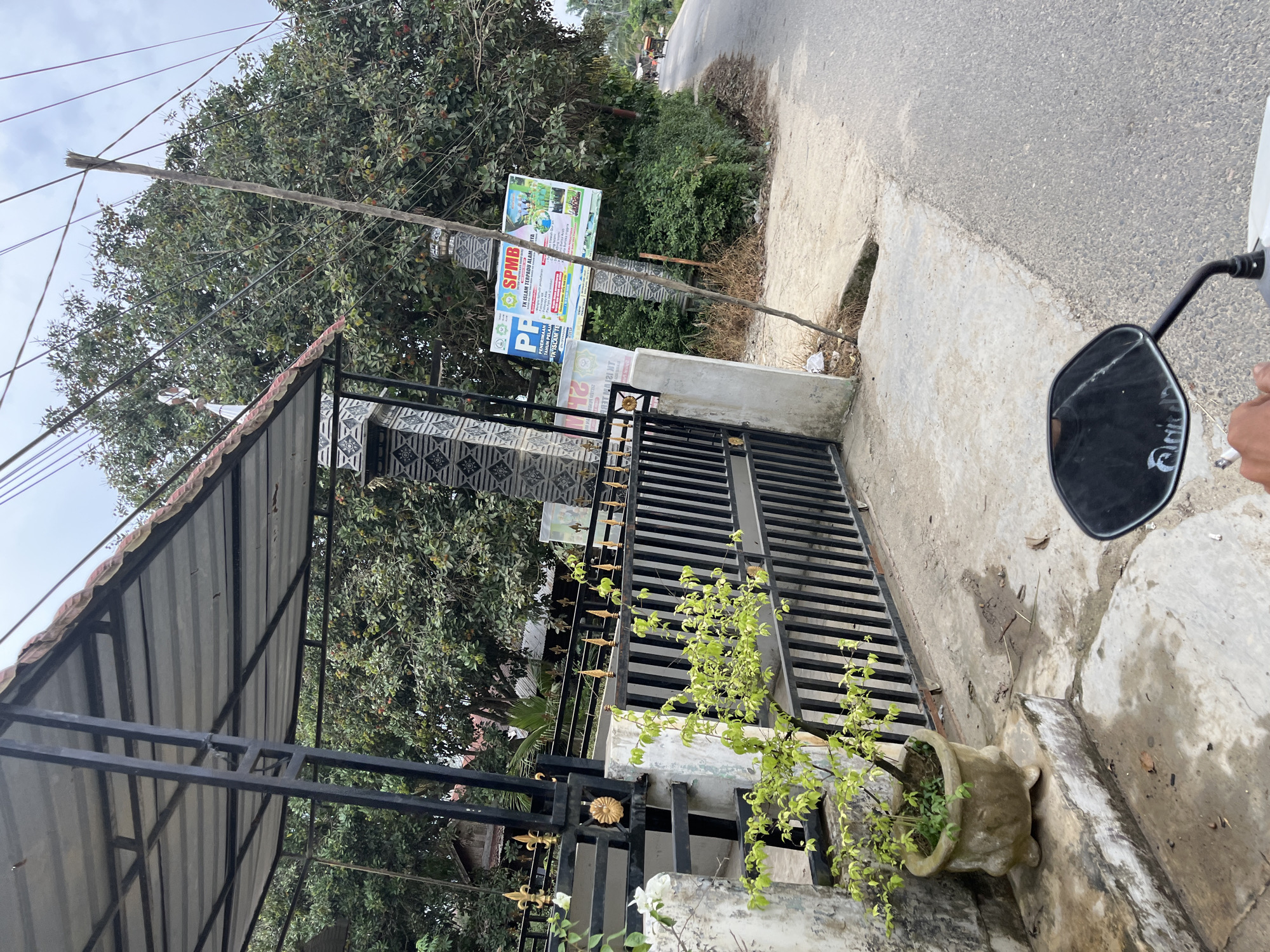 The image depicts a street-level view of a residential area or school entrance in Tanahterban, Indonesia, captured from the perspective of someone riding a motorcycle. In the foreground, the handlebar and a portion of a white motorcycle are visible, along with a black rearview mirror reflecting the sky and trees. A hand holding a cigarette is also present.
The focal point of the scene is a black metal gate with decorative gold accents, opening onto a concrete pathway. To the left of the gate, a potted plant with vibrant green leaves sits on a weathered concrete step. The gate is set against a white concrete pillar adorned with patterned tiles.
Behind the gate, lush green trees dominate the background, some bearing visible fruit that appears to be rambutan. A large sign with Indonesian text and colorful graphics, likely advertising a school or educational program, is affixed to a wooden pole amidst the foliage. The sign's text includes "SPMB TK ISLAM TERPADU ALAM" and "PENERIMAAN TAHUN PELAJAR".
The street itself is paved and appears to be a two-lane road with a slight incline. A few distant figures on bicycles or motorcycles can be seen further down the road, suggesting a moderate level of activity. The sky is overcast, contributing to the overall subdued lighting of the scene. The environment appears to be rural or semi-urban, characterized by dense vegetation and modest structures.