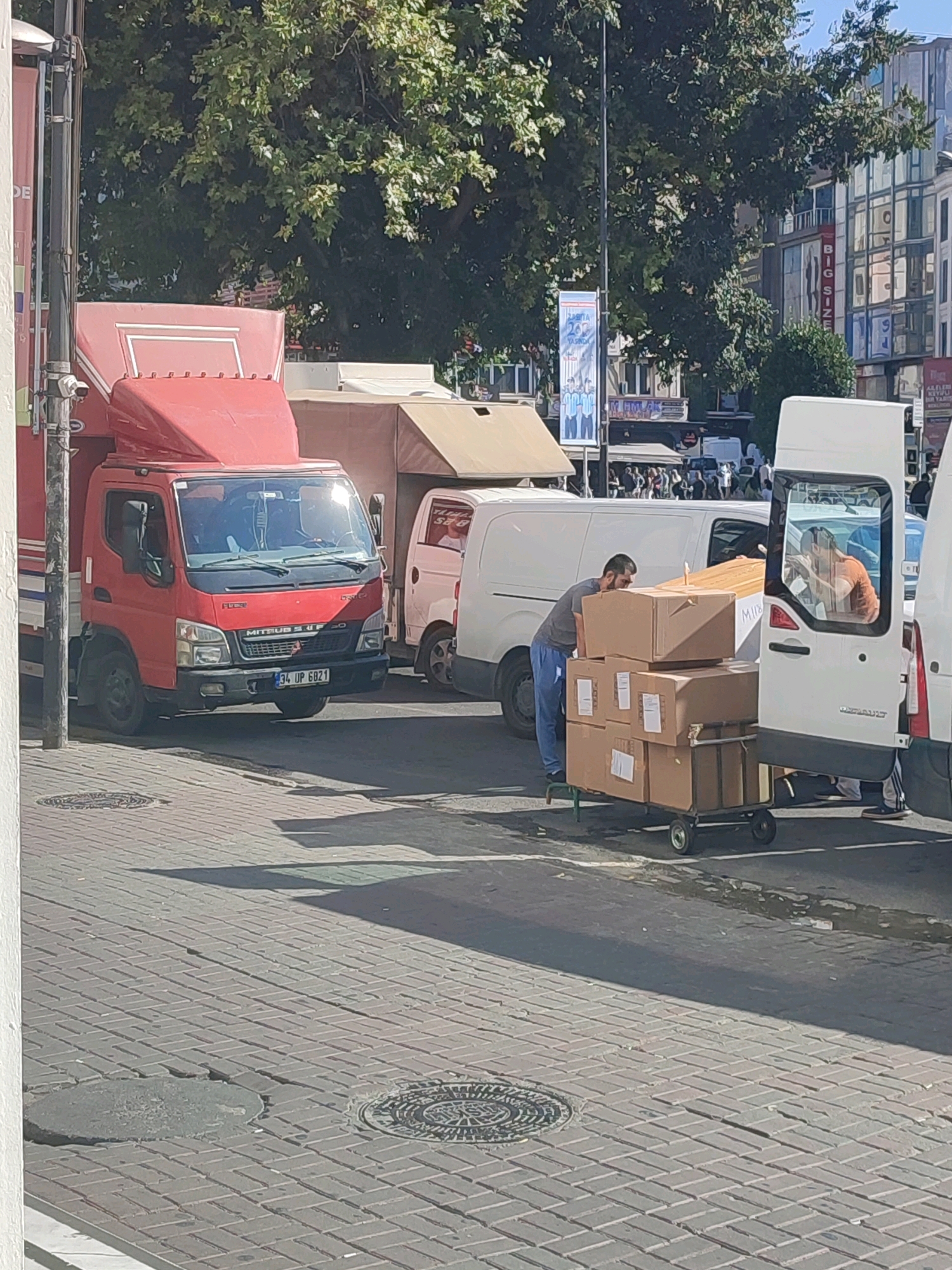 In Istanbul, Turkey, on a sunny day, a red delivery truck is parked on a cobblestone street. Beside it, a man wearing blue pants and a gray shirt is loading boxes onto a hand truck. The boxes are brown cardboard, some stacked precariously high. A white van is parked next to the hand truck, and a person with an orange shirt is visible through its rear window. In the background, trees and buildings line the street, suggesting an urban setting. The scene appears to be a busy delivery area.